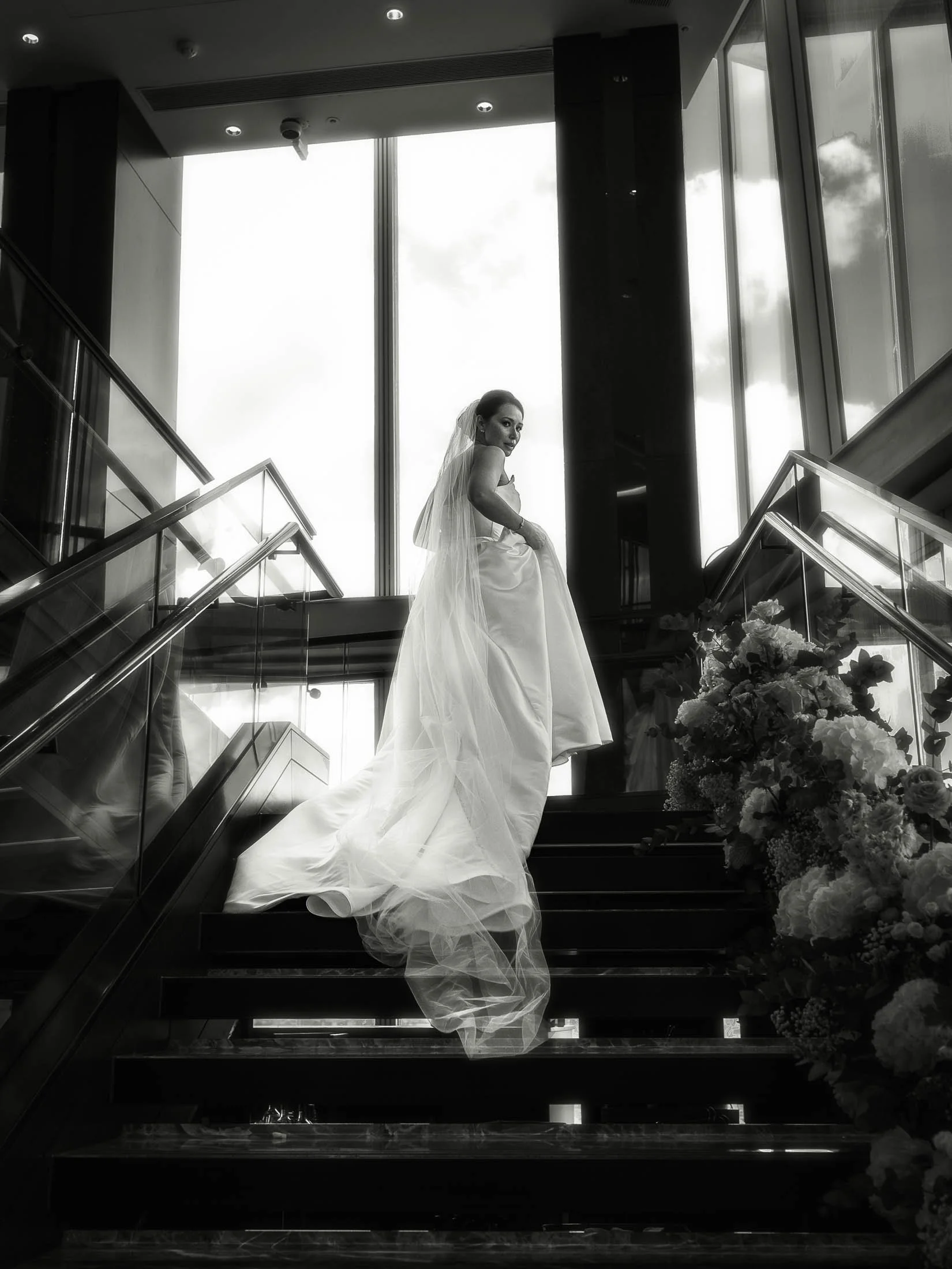An editorial bridal portrait in black and white, as an Australian bride walks up the staircase at the Shangri-La hotel in The Shard, London, holding her luxurious wedding dress and looking back at the camera.