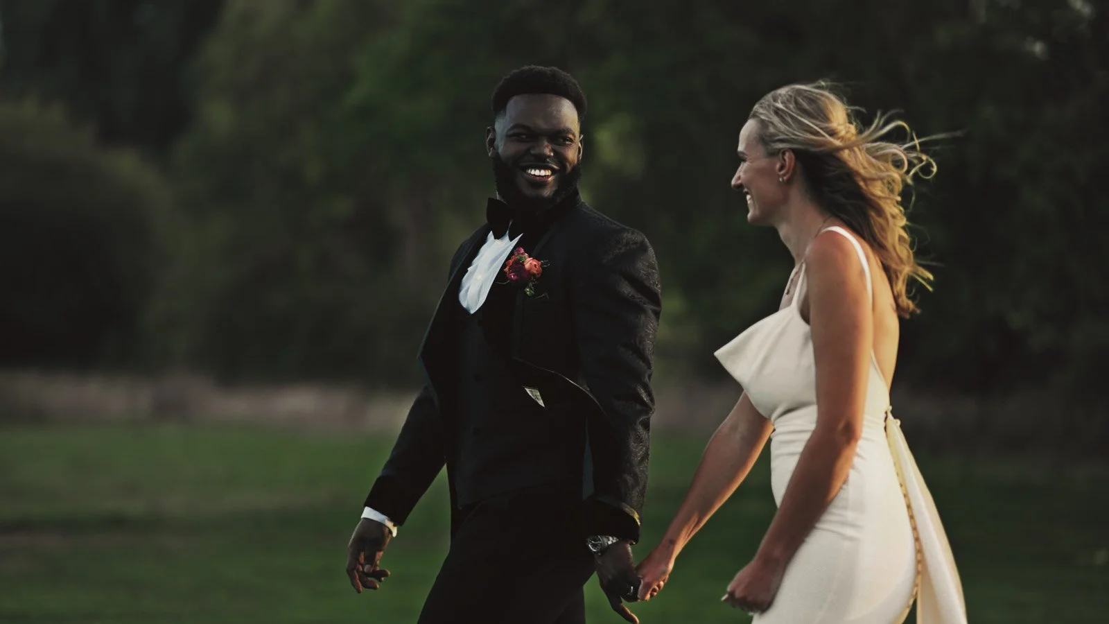 A couple walking along a path on the banks of the River Thames during their colourful riverside wedding. The groom is wearing black tie and the bride is wearing a simple, elegant gown.