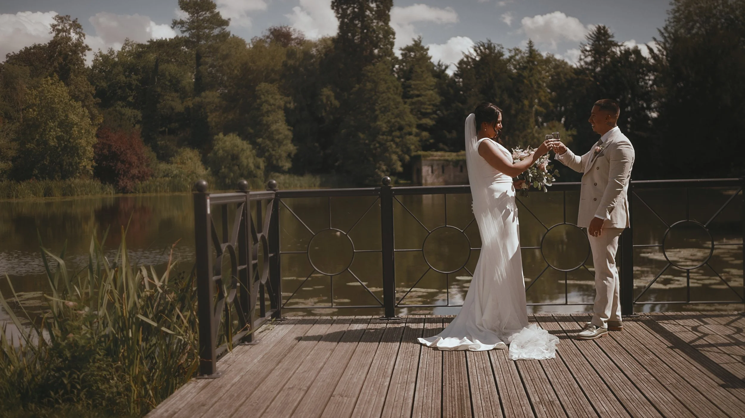 A couple toast each other with Champagne by the lake during their outdoor wedding day at The orangery in kent.
