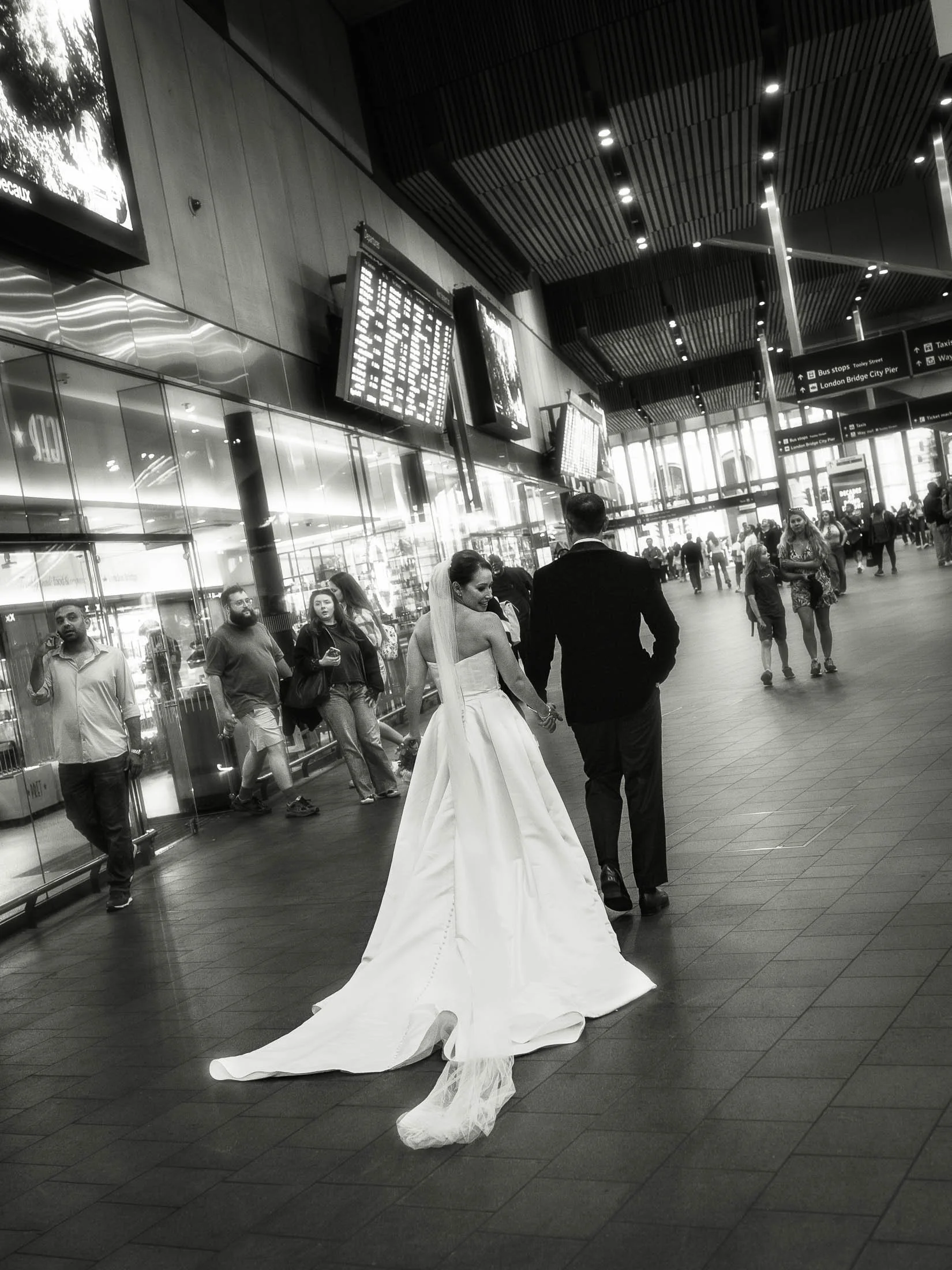 A romantic and editorial moment during a black tie London wedding as the bride and groom hold hands and walk through the London Bridge station concourse. Captured in a black and white timeless aesthetic.