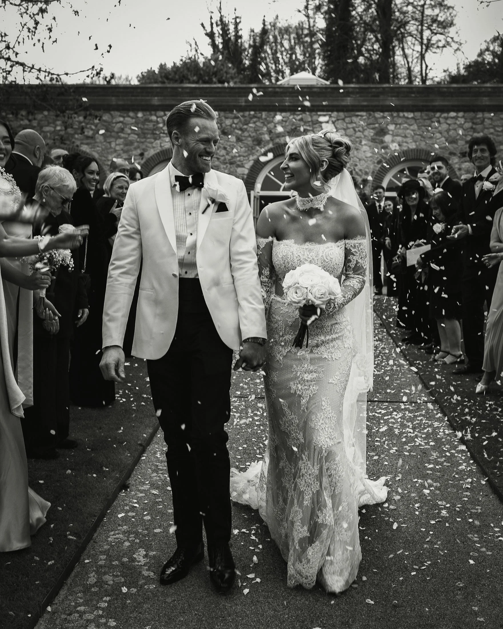 Black and white editorial wedding moment of a bride and groom in white tuxedo walking through confetti and smiling at The Orangery Maidstone.