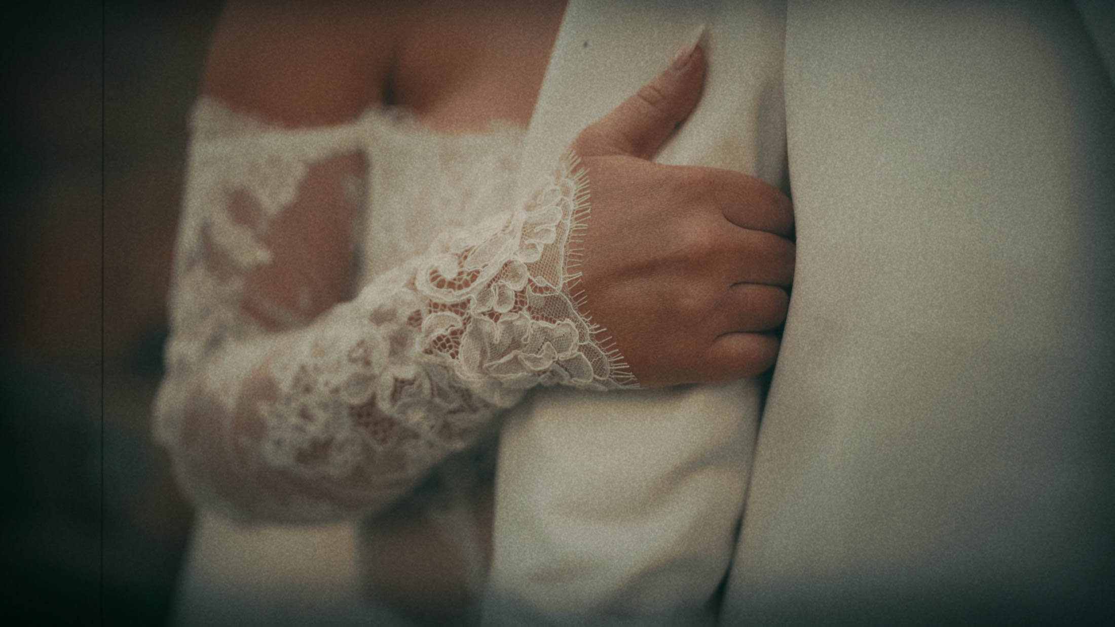 A brides hand holds onto the arm of her groom in white tuxedo, showing the detailing on her wedding dress. Captured in a super 8mm emulation style as part of the intro from a storytelling wedding film.
