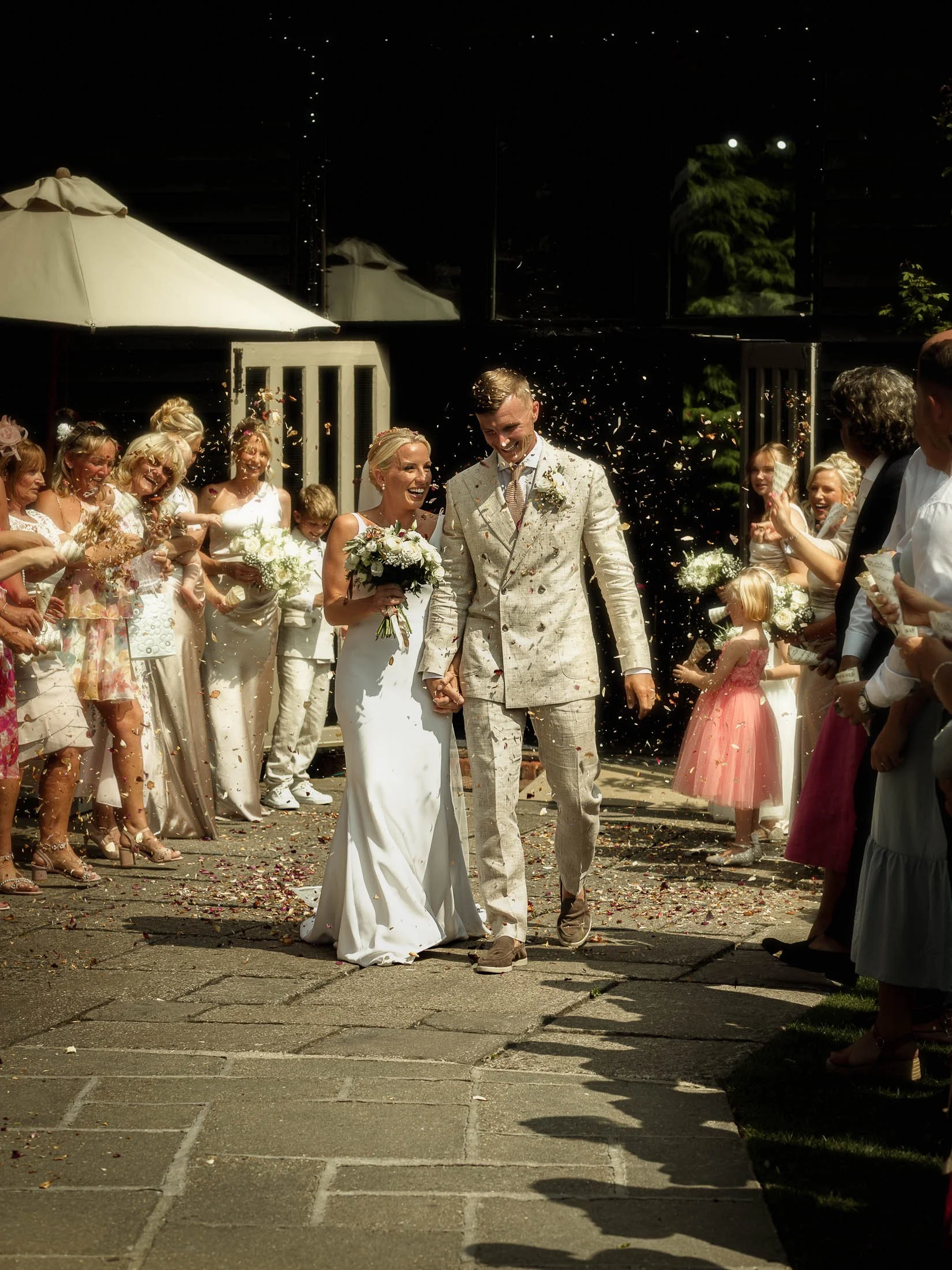 A happy wedding photograph showing a couple laughing together as they exit their outdoor wedding ceremony under a shower of confetti and being cheered by their wedding guests. Captured during a colourful outdoor wedding at Winters Barns in Kent, UK 