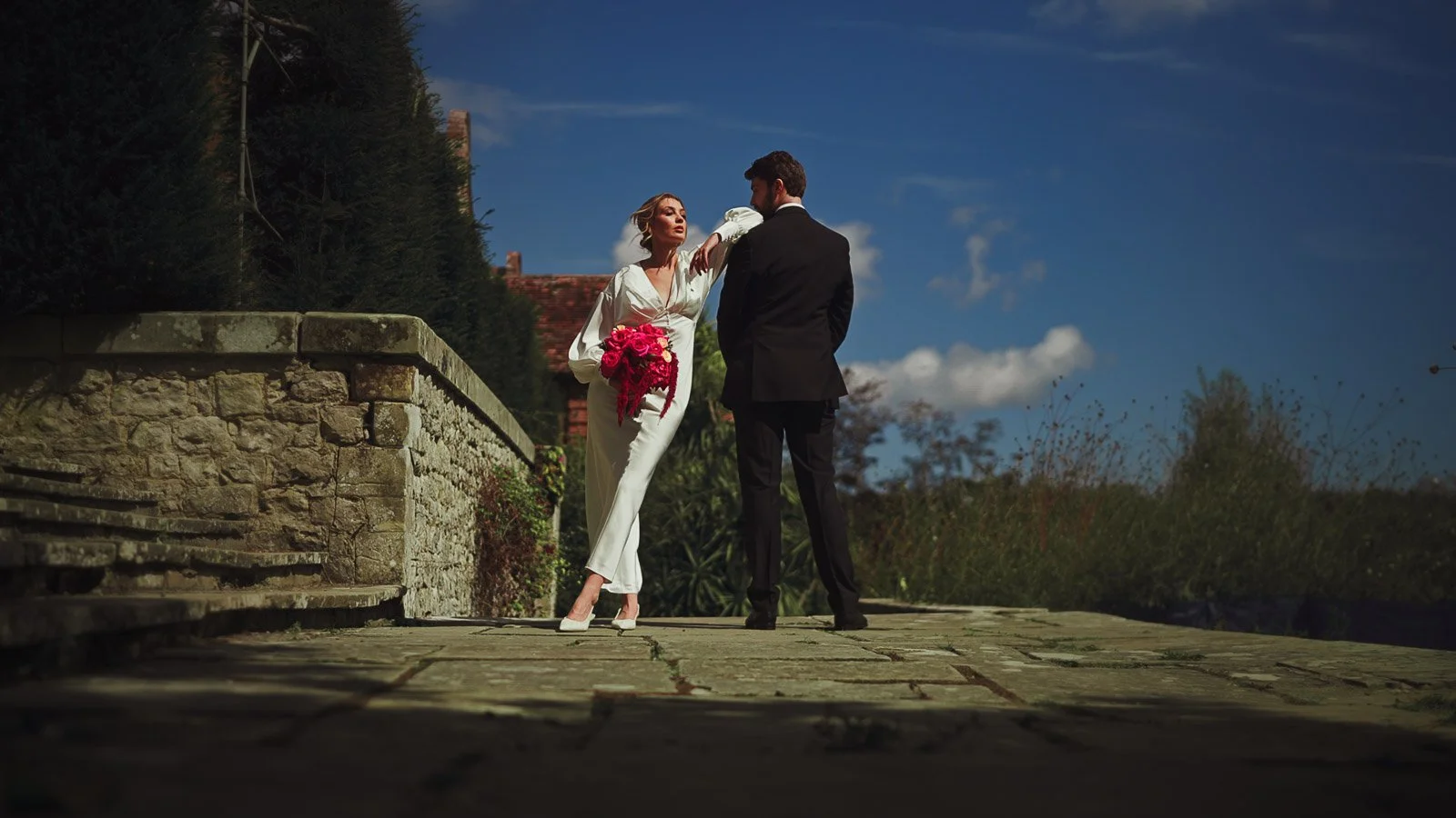 A fashion-focussed moment between a bride and groom standing on a veranda with bright blue sky behind them.
