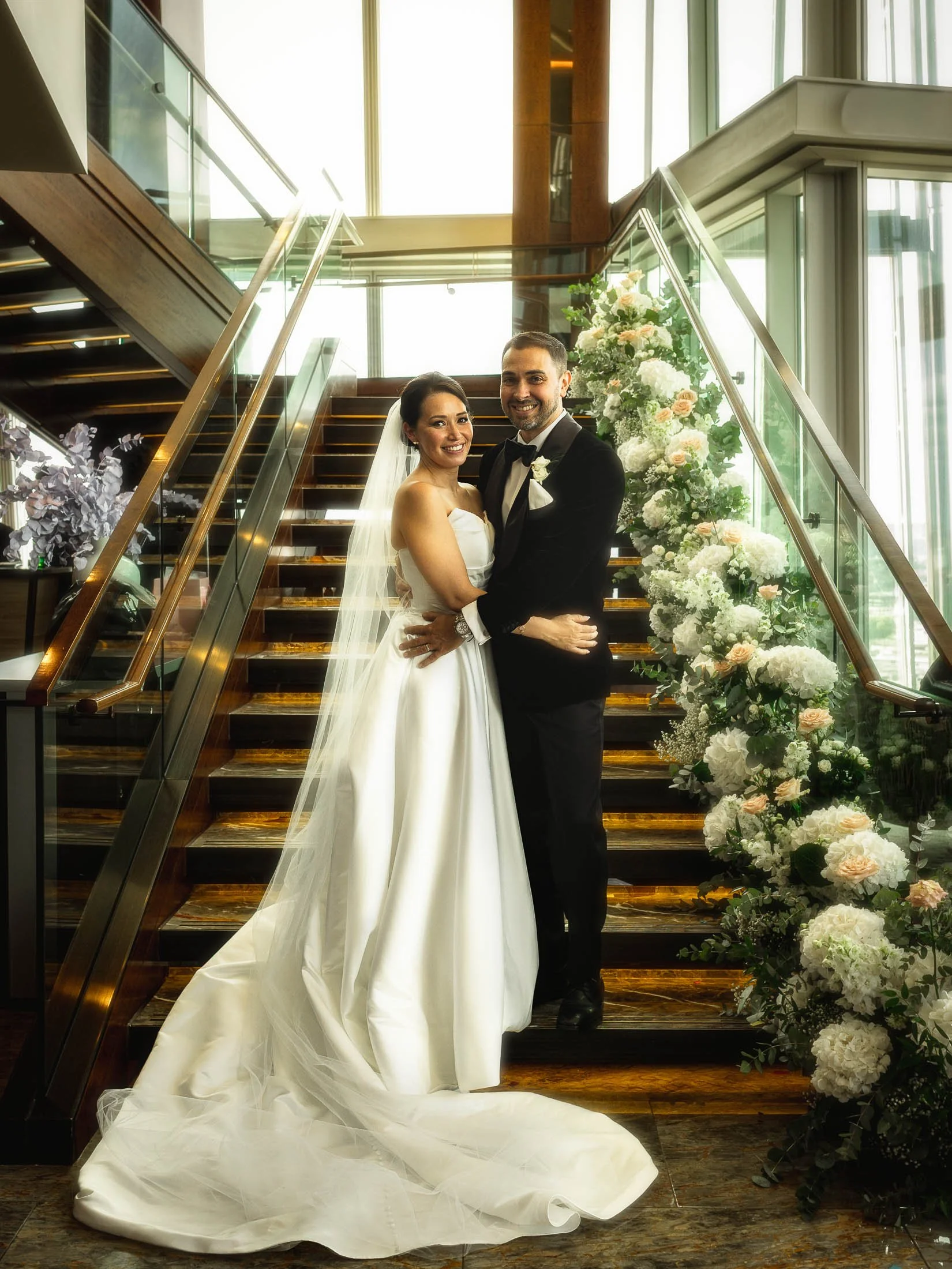 A formal picture of a bride and groom smiling at the camera as they are captured at the bottom of the staircase on the 34th floor of the Shangri-La The Shard hotel during their black tie London wedding.