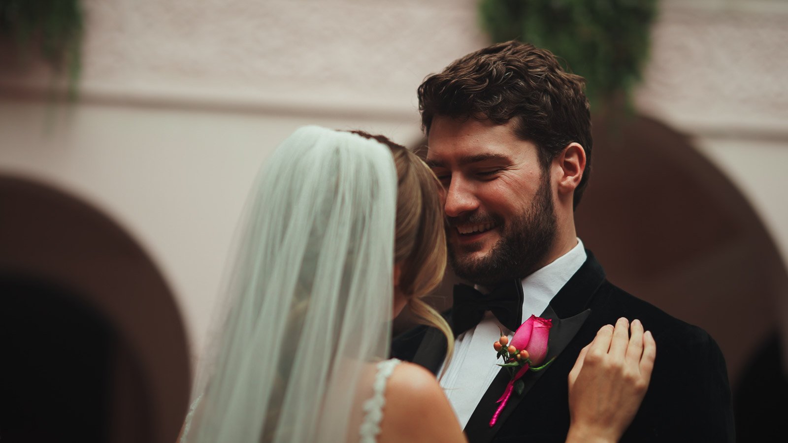 Groom in velvet jacket and black tie with bright pink button hole dances with his bride.