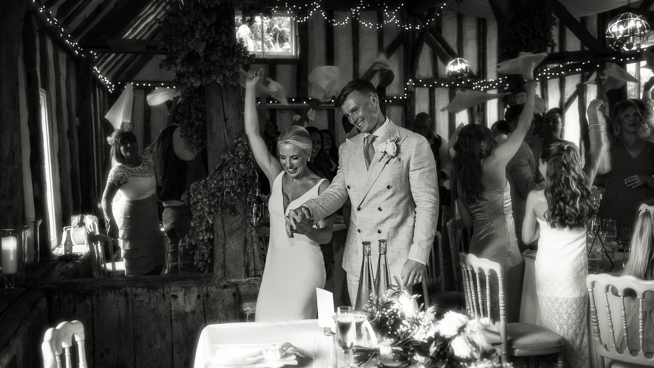 A bride and groom enter their wedding reception at Winters Barns in Kent to cheers and napkin waving by their guests. Captured by both wedding photography and videography as part of a cohesive team approach.