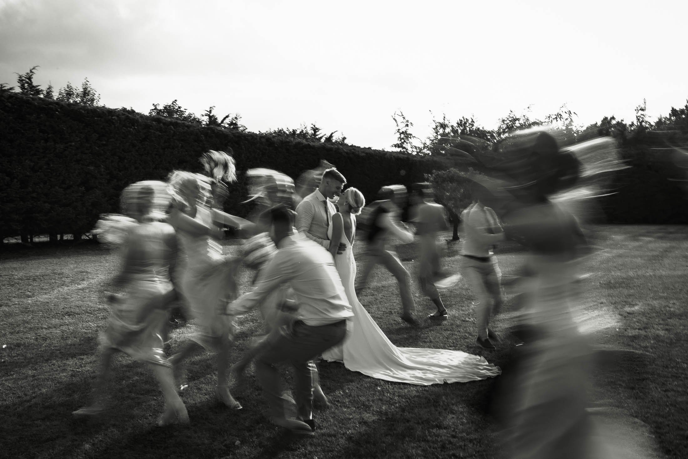 An editorial and artistic wedding photograph showing a shutter drag effect, as the bride and groom are in focus in the centre as their bridal party are blurred running around them. A black and white image captured at Winters Barns in Kent.
