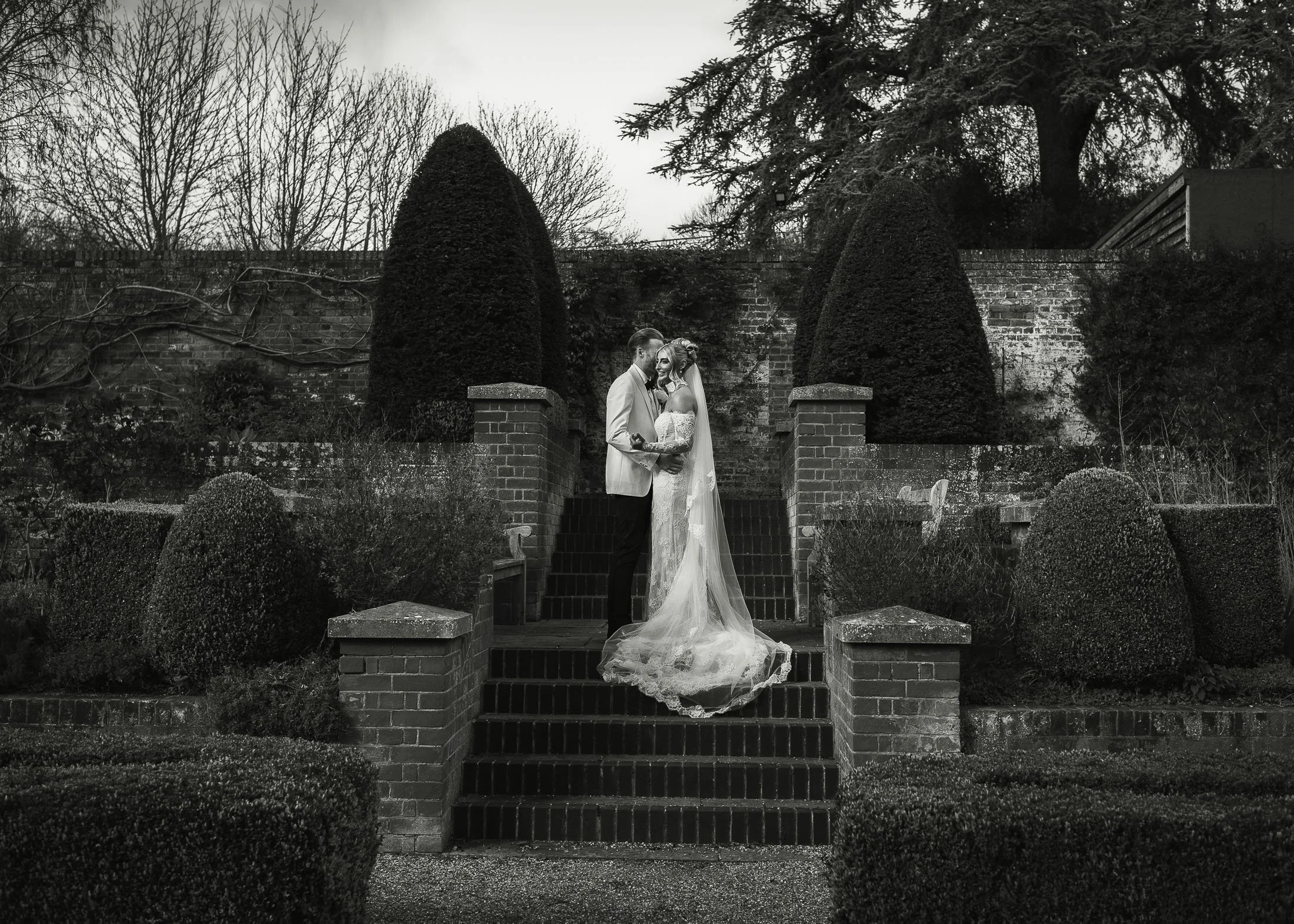A groom wearing a white tuxedo embraces his bride on the steps of the formal gardens during a wedding at The Orangery Maidstone.