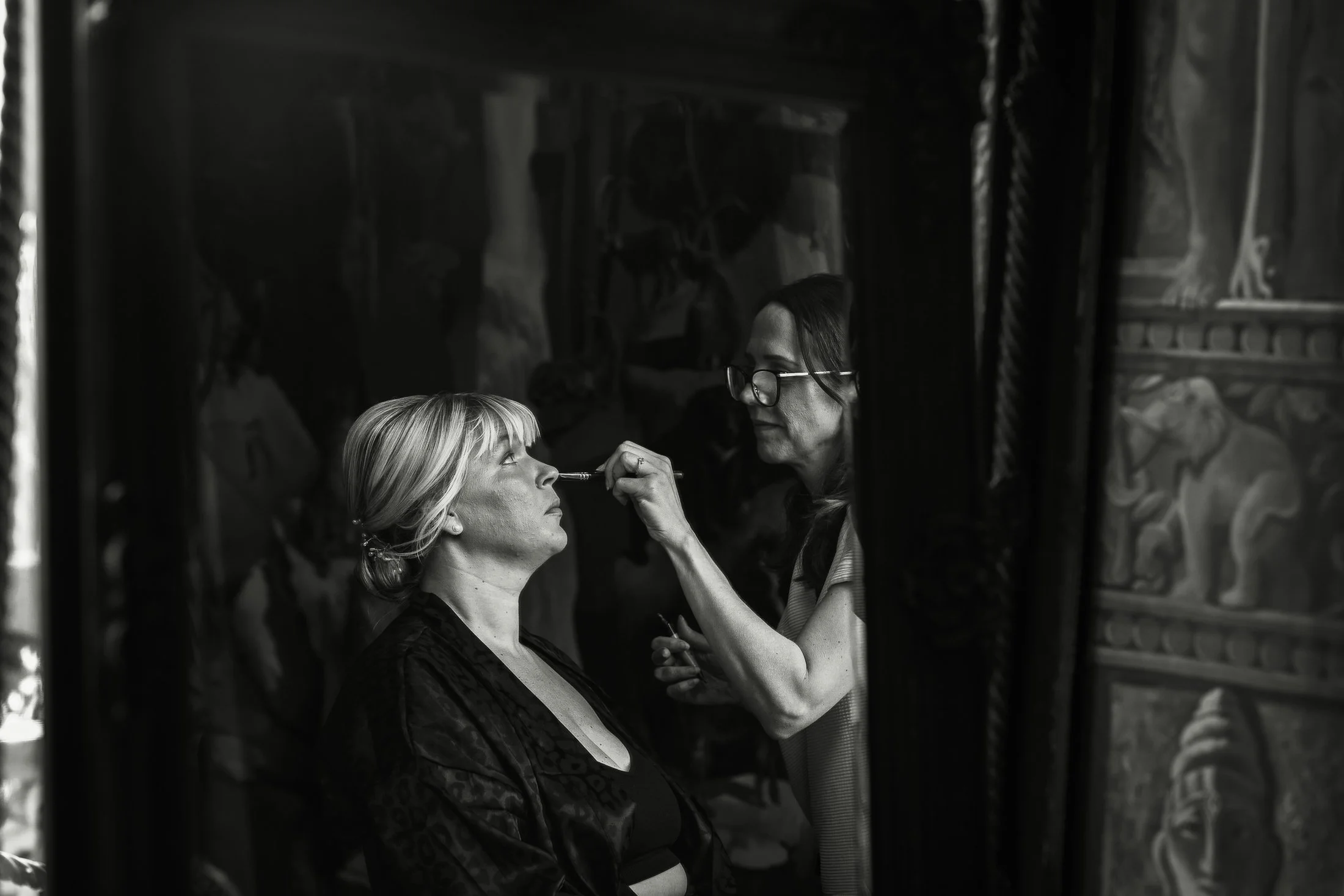 A reflection in a full length mirror of a bridesmaid having her make up applied.