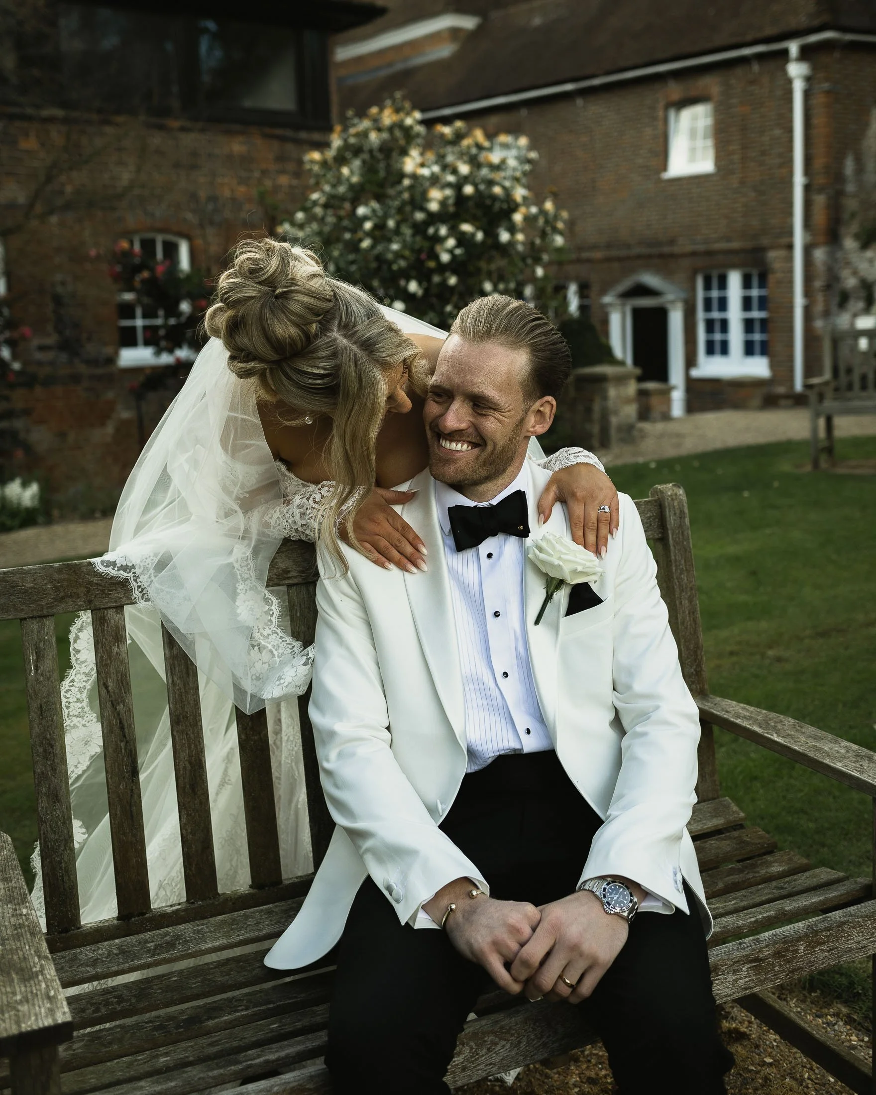 A groom in white tuxedo sits on a bench as his bride leans on him and smiles at The Orangery Maidstone.