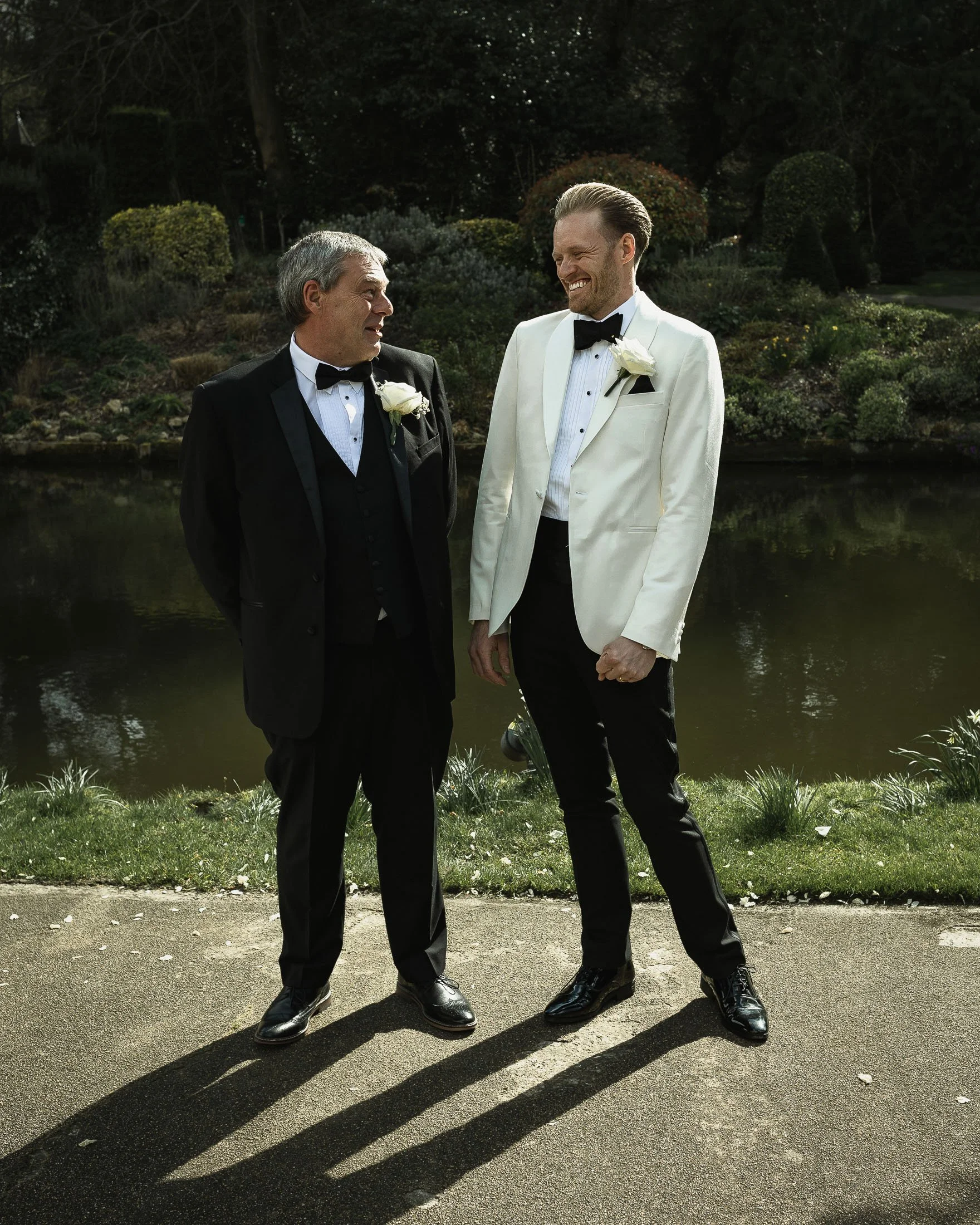 Formal group shot of a groom in white tuxedo with his father by the lake at The Orangery Maidstone wedding venue.