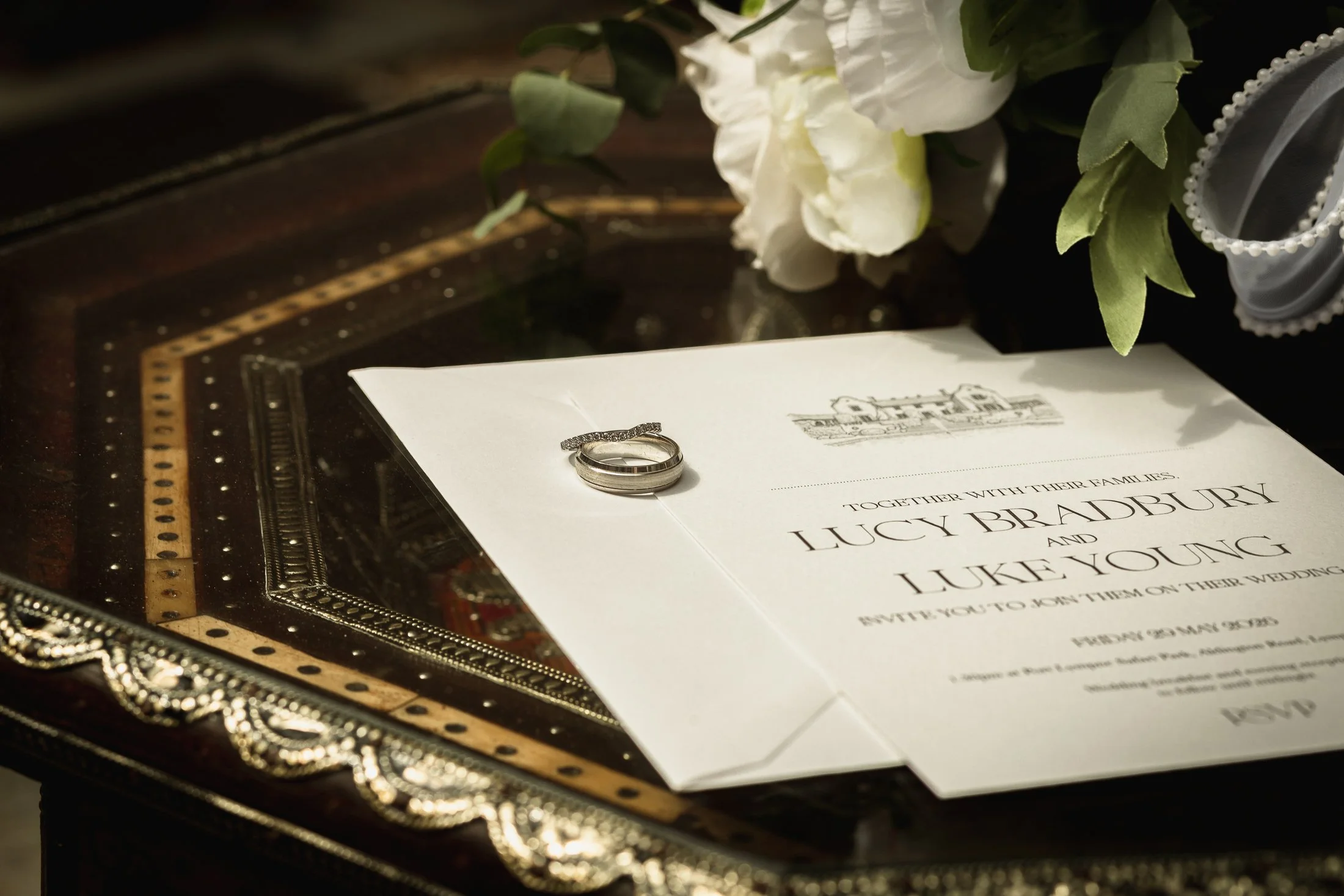 Lucy & Luke's wedding rings sit on top of a wedding invitation, next to a bouquet of white flowers in the Moroccan Courtyard at Port Lympne.