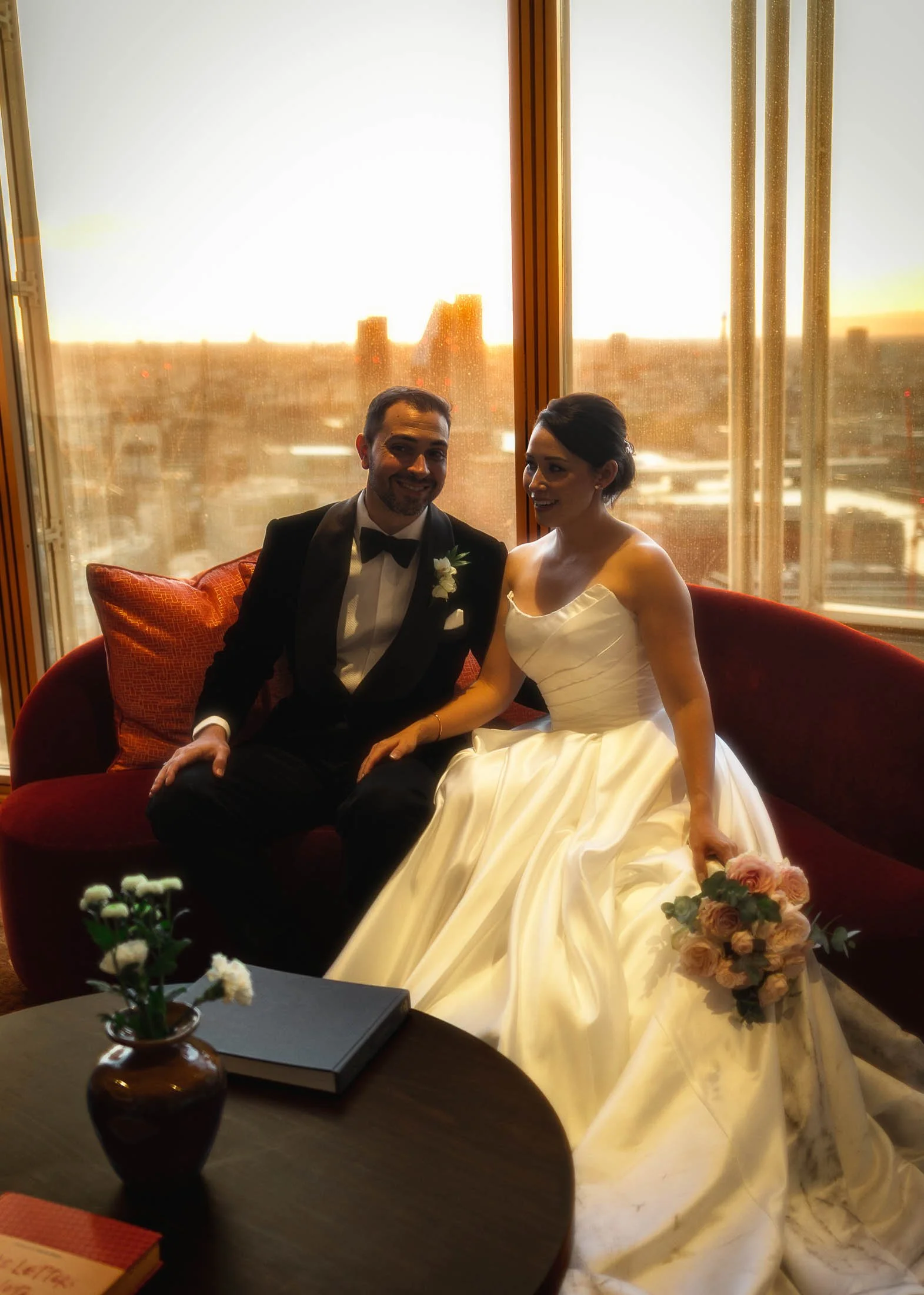 A bride and groom sit on a red sofa at the Shangri-La The Shard hotel during golden hour on their romantic and international black tie wedding day.
