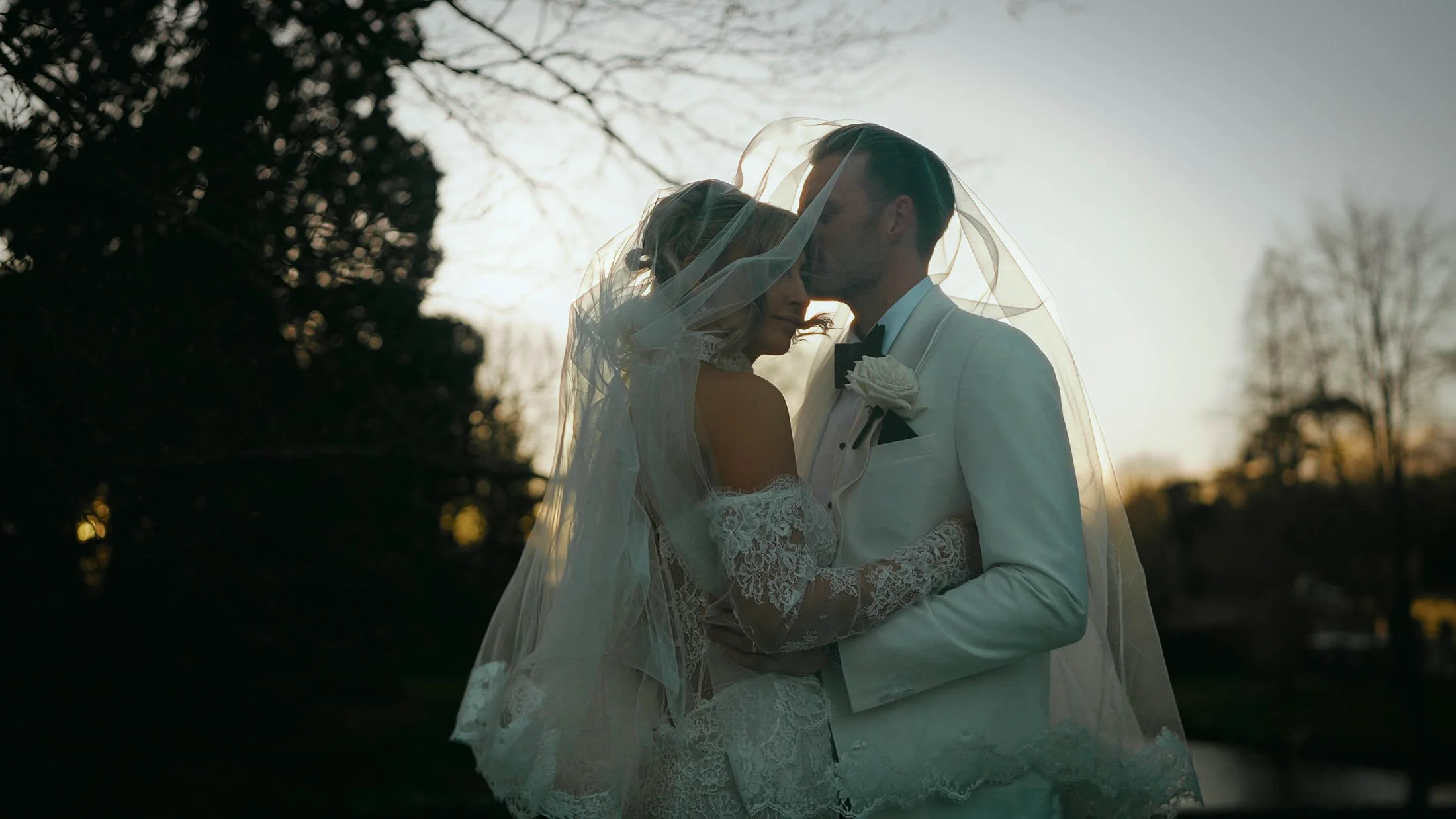 A groom kisses the forehead of his bride as they stand underneath a veil at sunset.