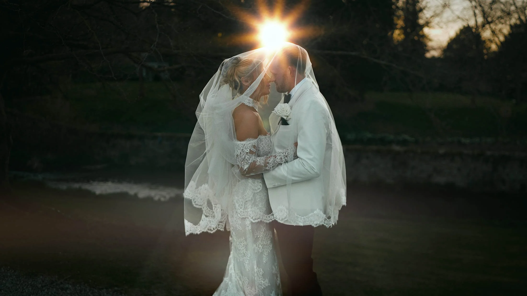 An iconic storytelling wedding film moment as the couple stand underneath the brides veil as the sun sets and sun flares appear across the frame.