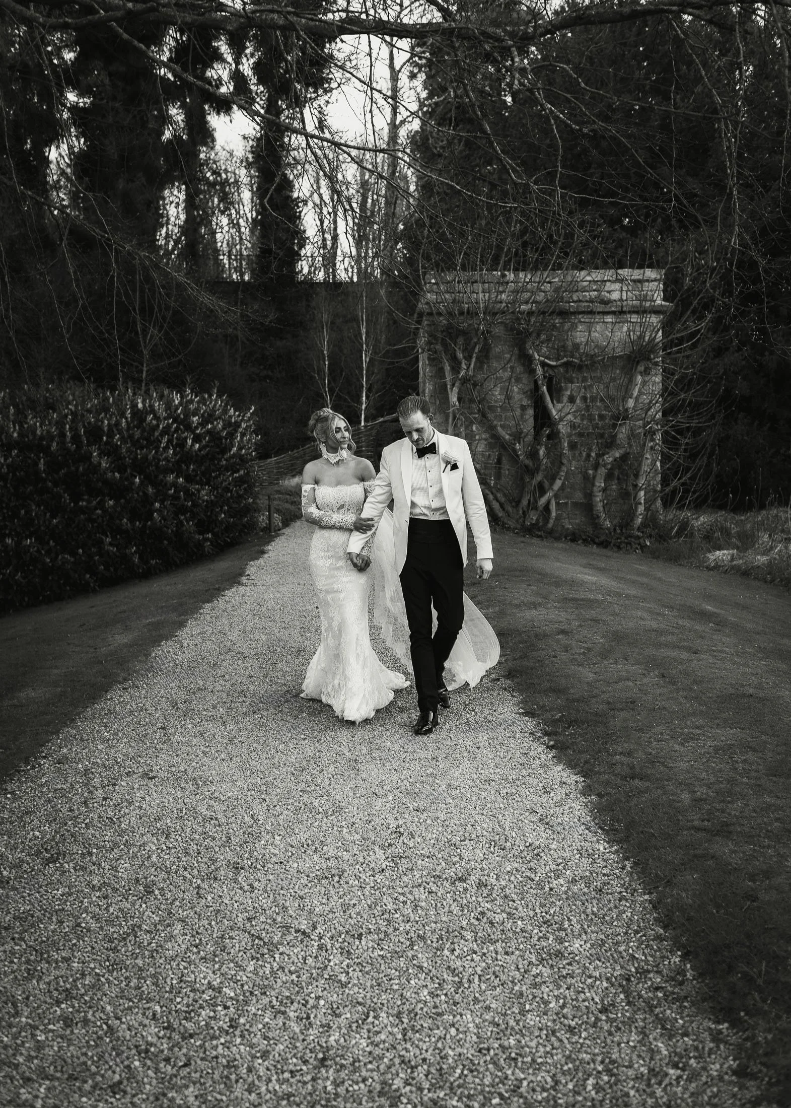 White tuxedo groom and bride walk along the gravel path by the lake at The Orangery Maidstone.