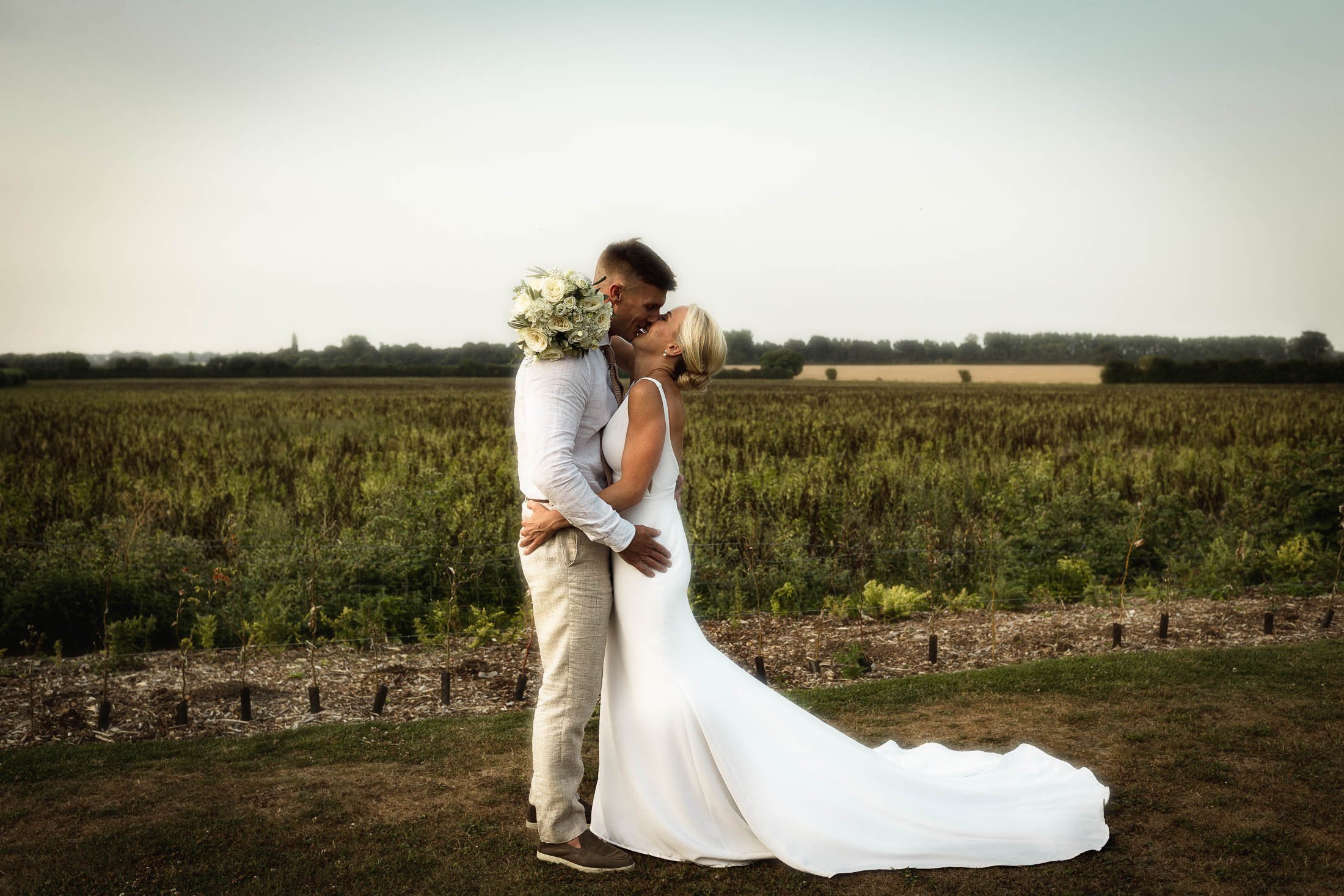An editorial wedding pose of a bride and groom as they embrace and laugh together, with the rolling Kent countryside behind them. Captured during a summer wedding day at Winters Barns.