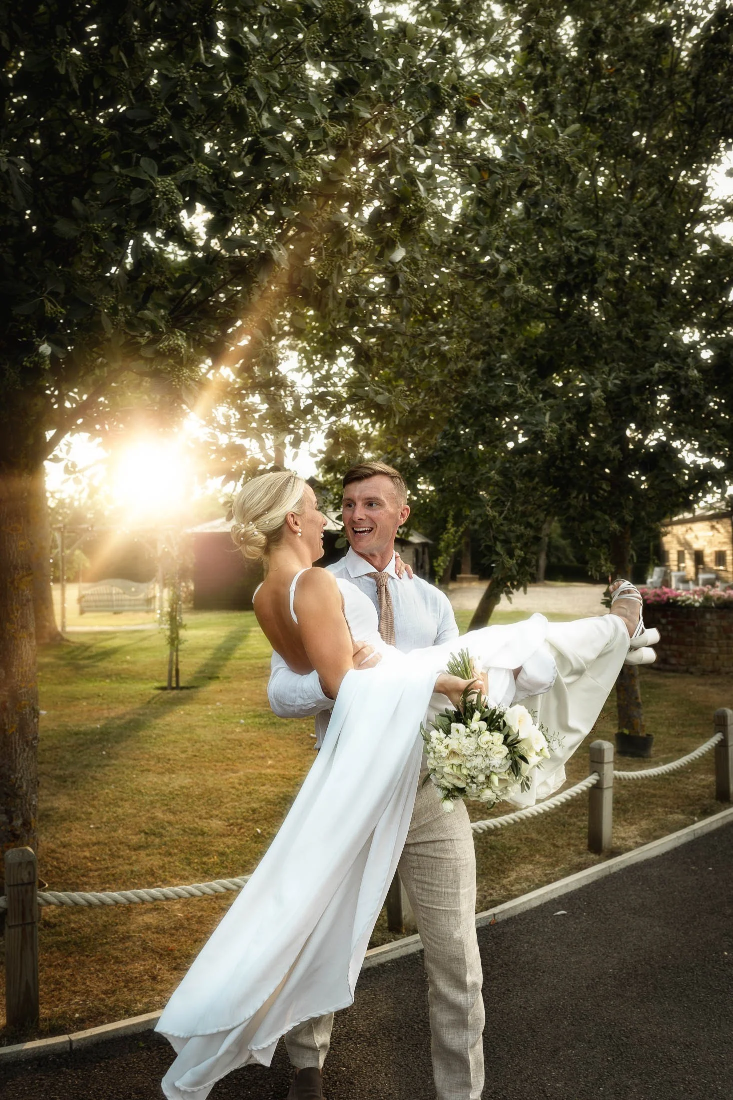 A groom picks up and carries his bride as golden hour sun flares come through the trees in the background, captured during a golden hour portrait session at Winters Barns.