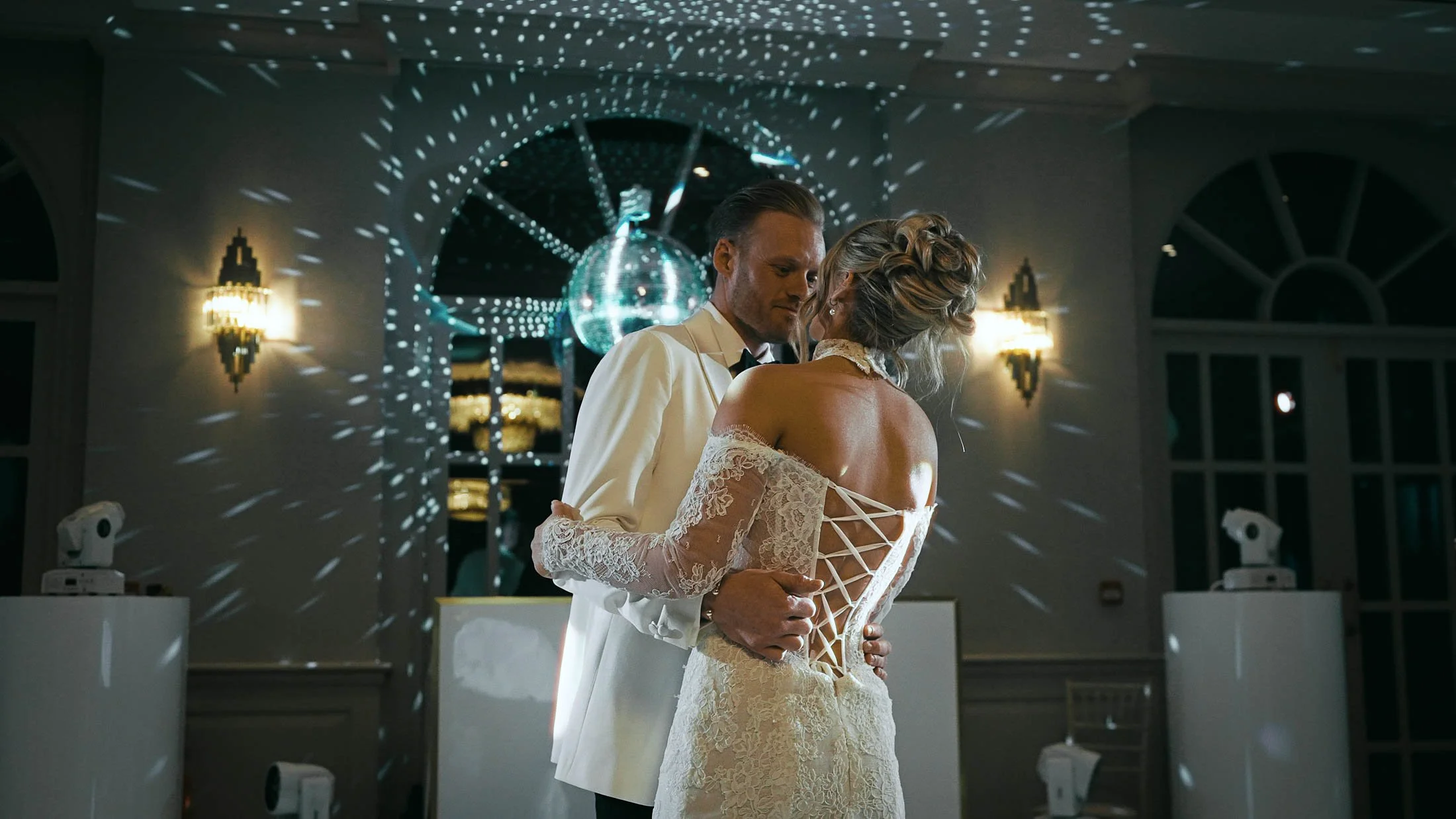 A couple share their first dance as a glitter ball reflects lights onto the wall behind them, in a reflective moment during their cinematic storytelling wedding film.