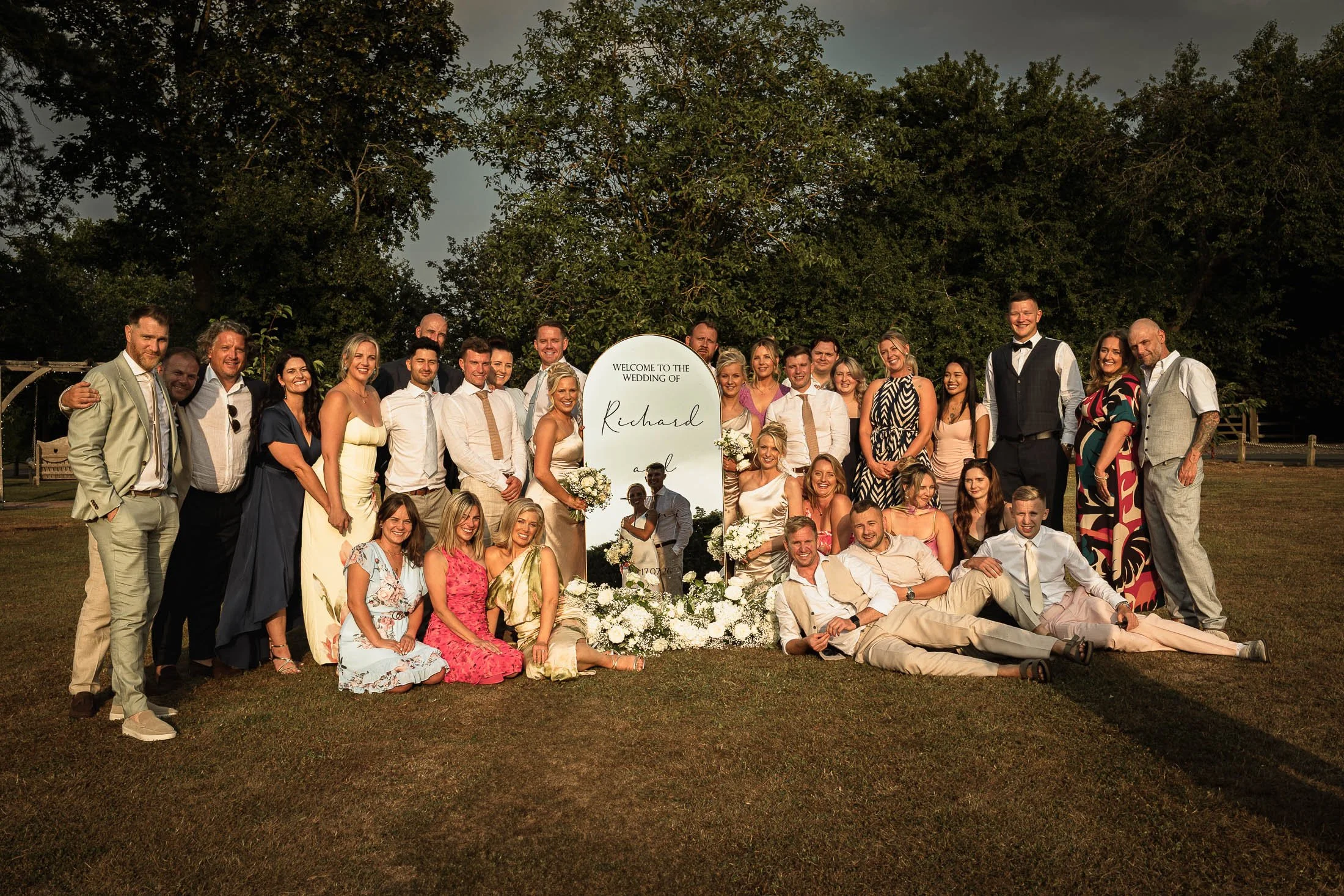 A bridal party surround a large mirror at sunset, with the reflection of the mirror showing the bride and groom. Captured in the grounds of Winters Barns wedding venue during sunset.