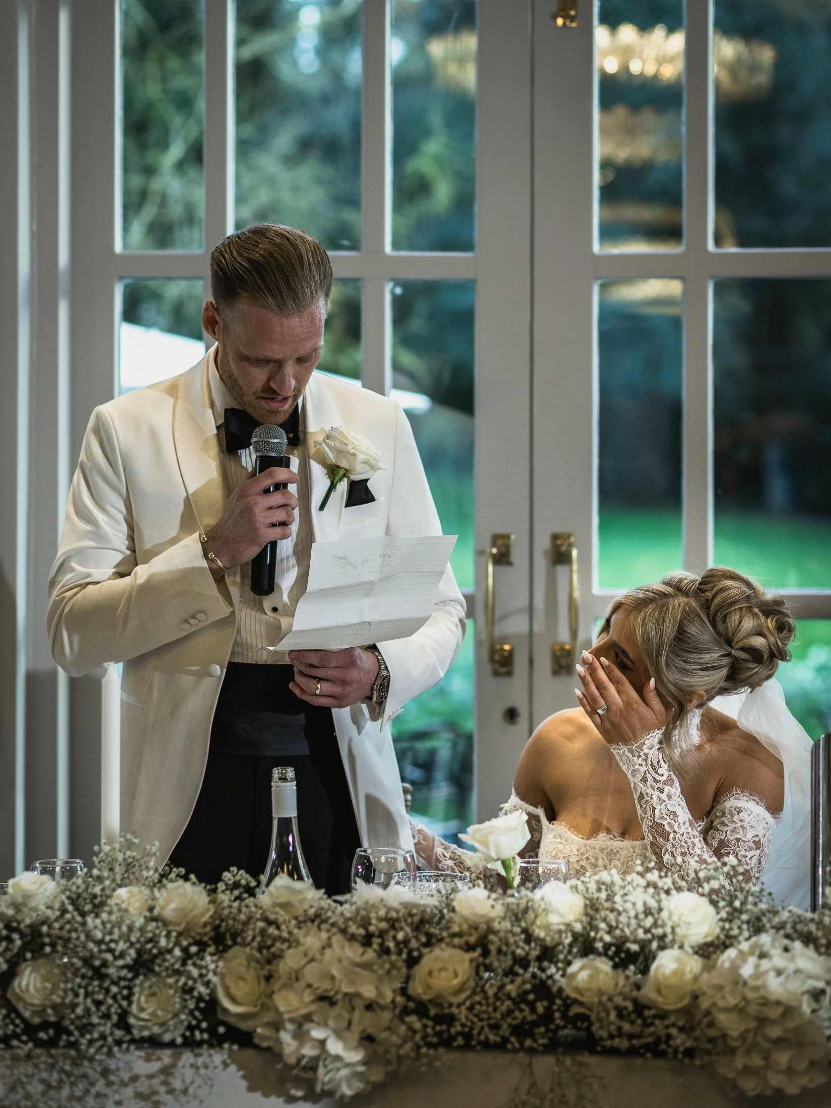A groom in white tuxedo delivers his speech as his bride wipes a tear from her eye.