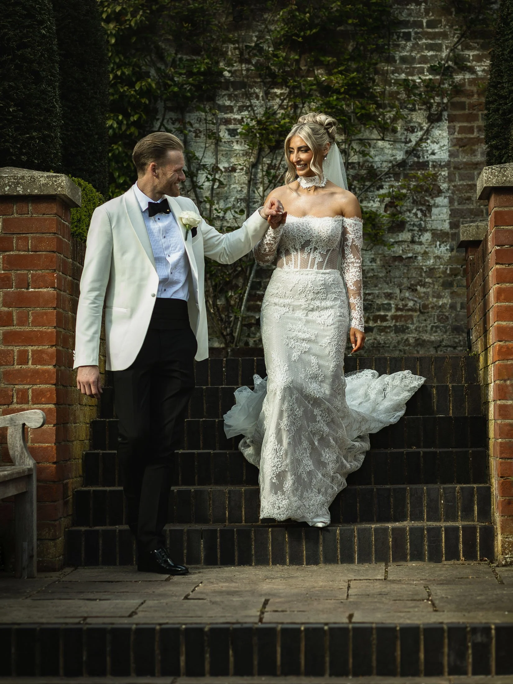 A groom in white tuxedo leads his bride down steps during their black-tie wedding at The orangery Maidstone.