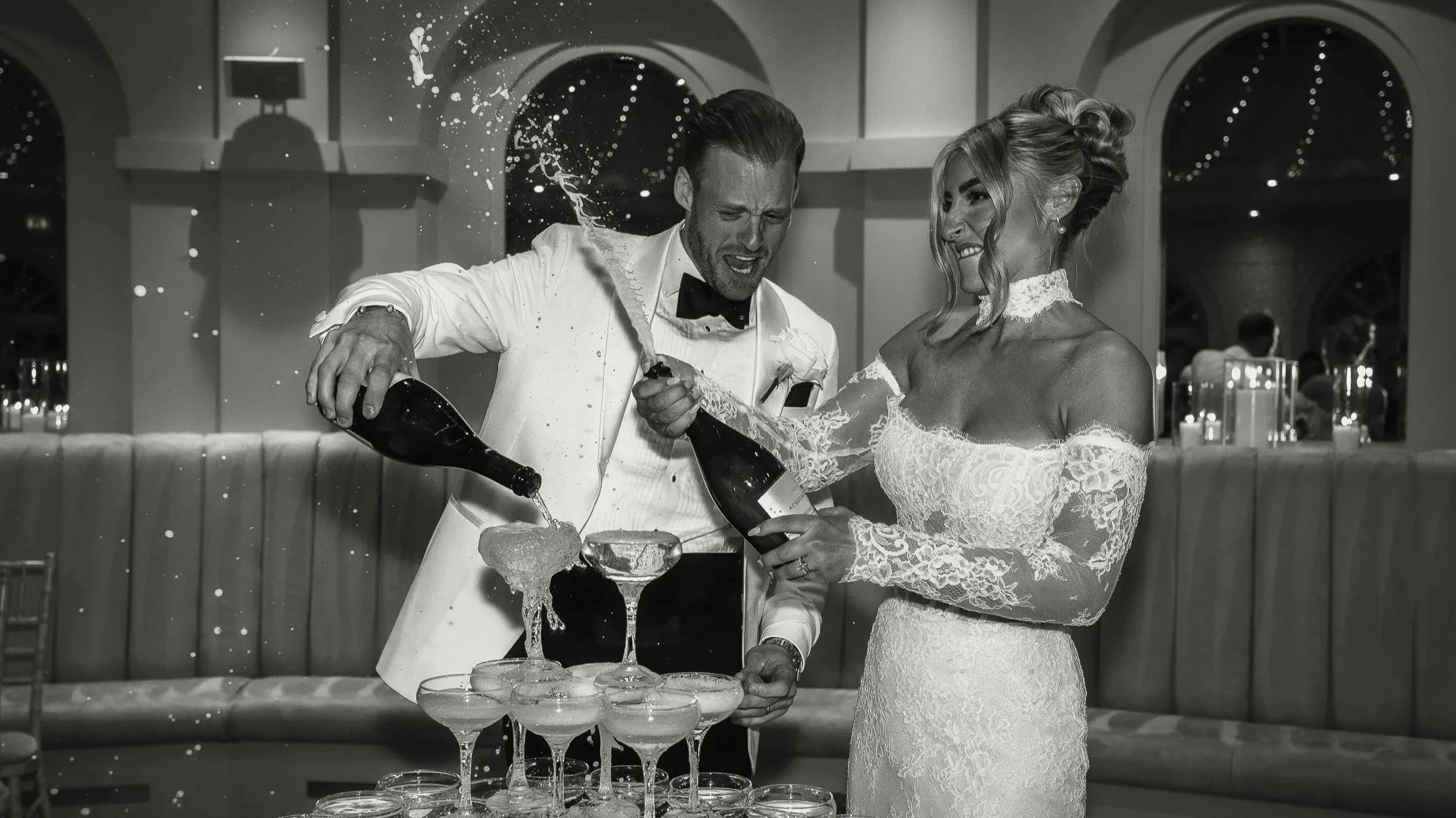Bride sprays Champagne from a bottle as the groom laughs.