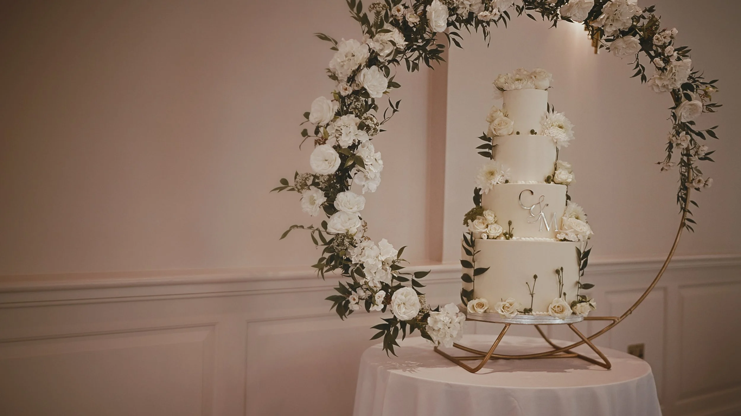 A luxury four tier wedding cake sits within a floral arch at a wedding at The orangery in kent.