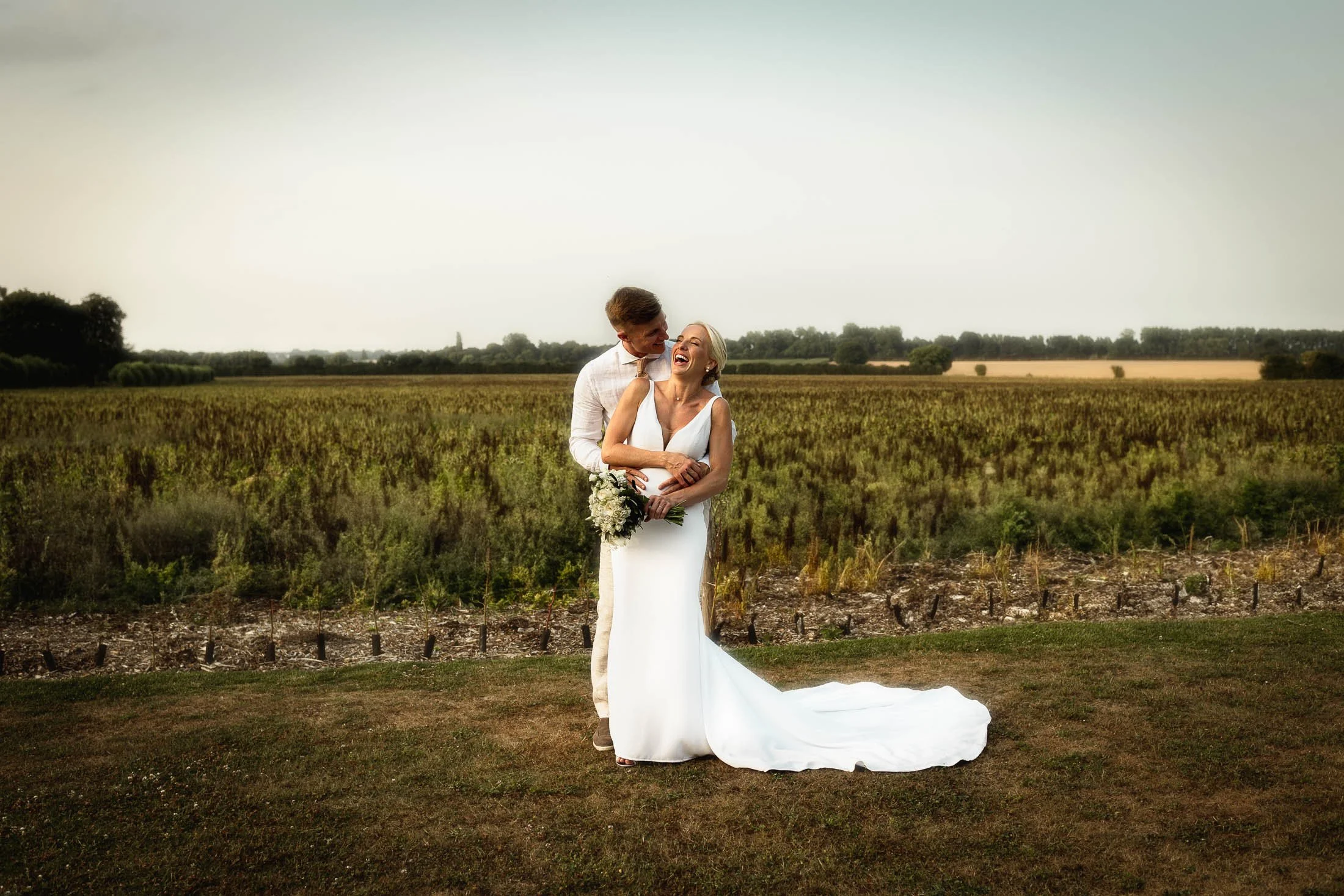 The groom wraps his arms around his bride as she laughs in a golden hour portrait captured during a summer wedding at Winters Barns in Canterbury, Kent.