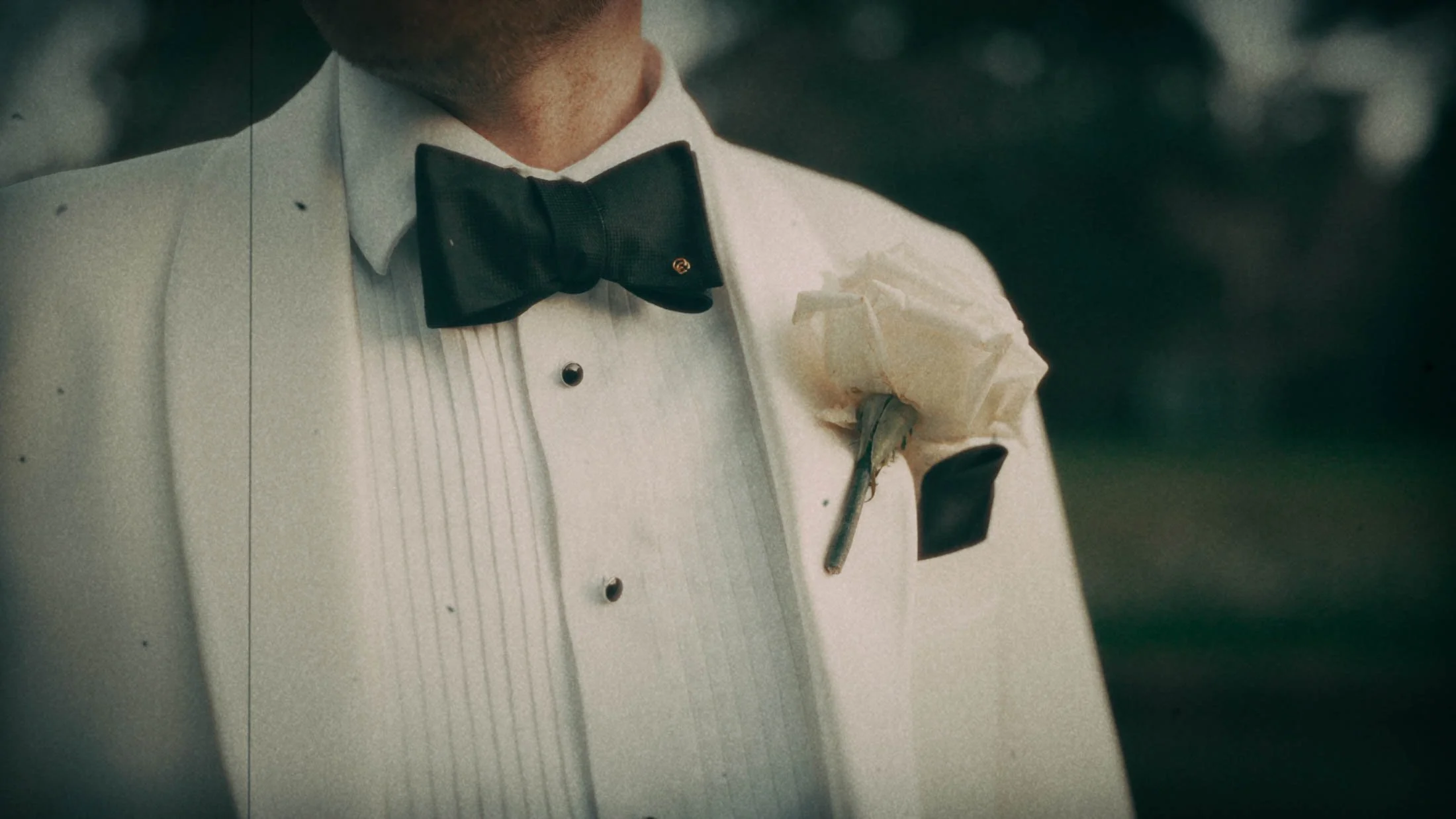 A detailed shot in a super 8mm emulation style of a Grooms white tuxedo jacket, bow tie and button hole from a storytelling wedding film into.