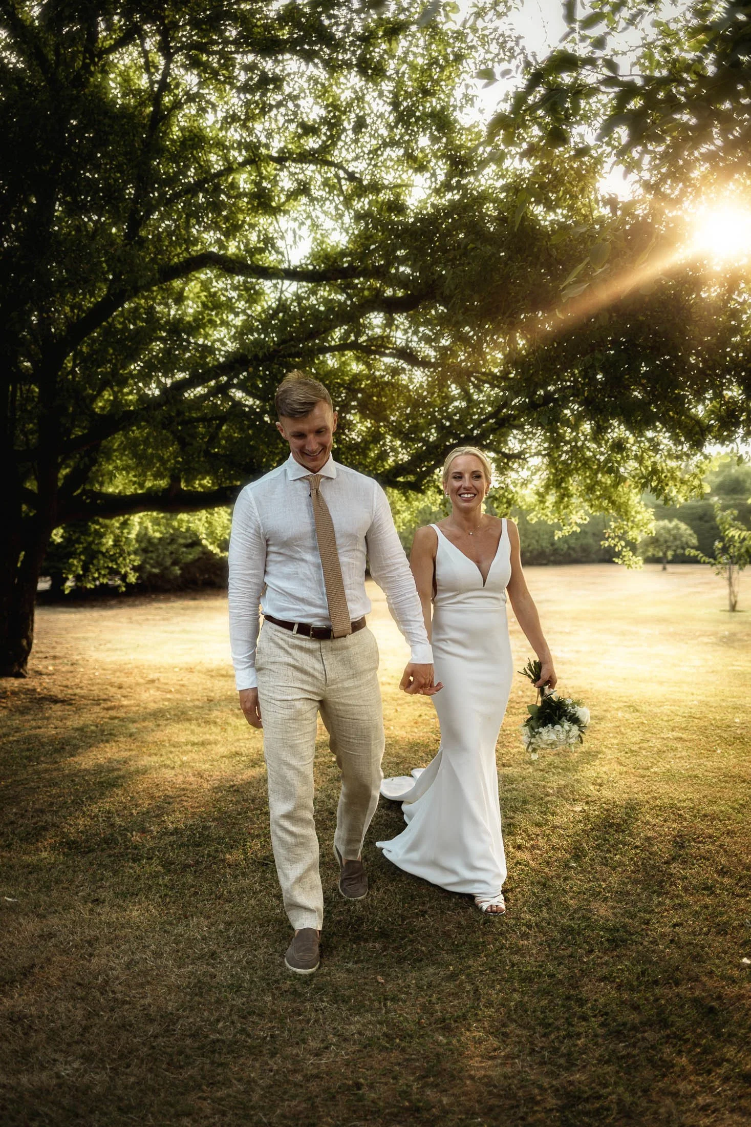 A bride and groom walking hand in hand underneath trees as the golden hour sunlight pours through the leaves, in an editorial portrait at Winters Barns wedding venue in Kent.