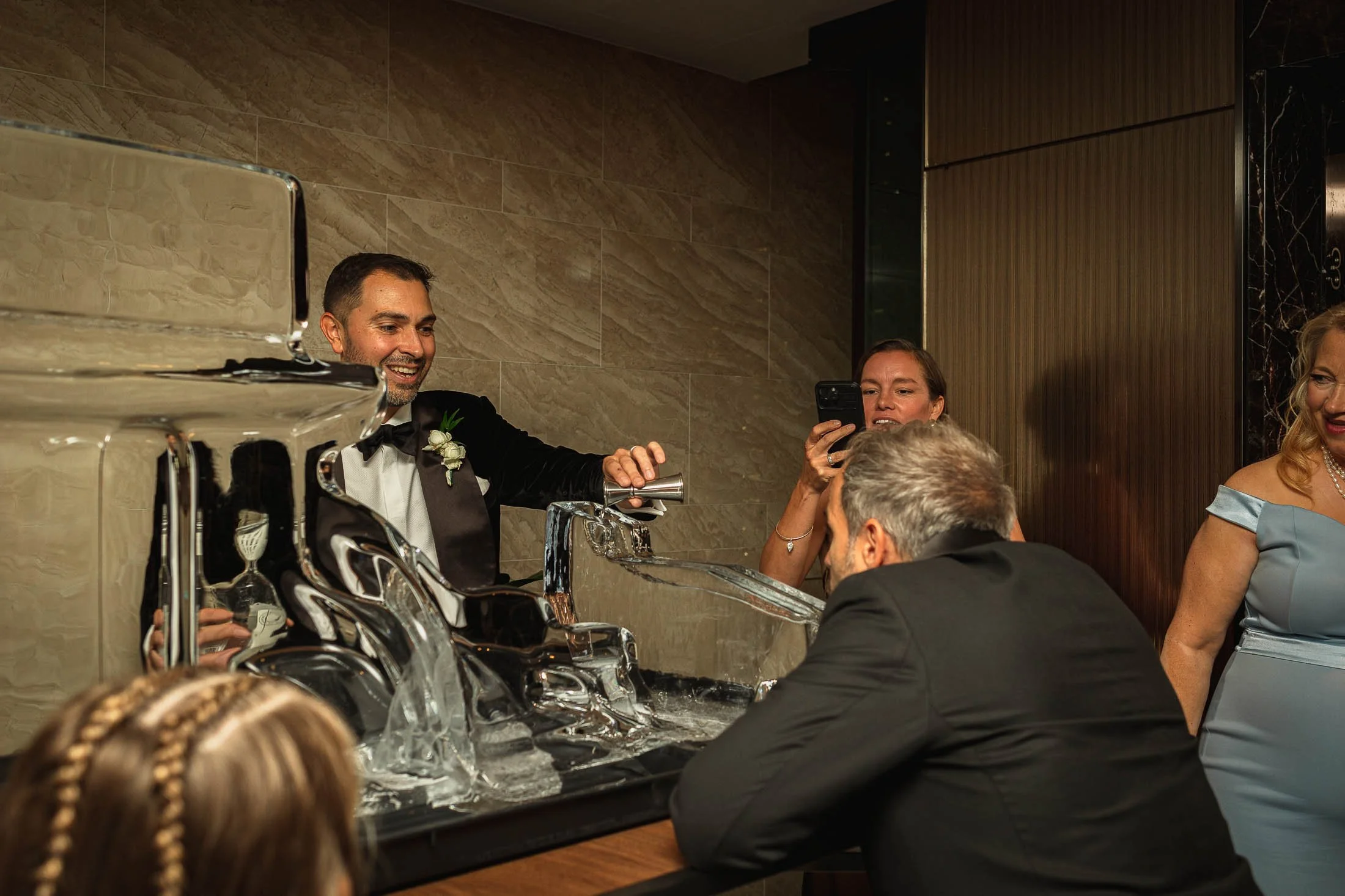 A groom in black tie tuxedo pours a drink into the mouth of one of his guests.