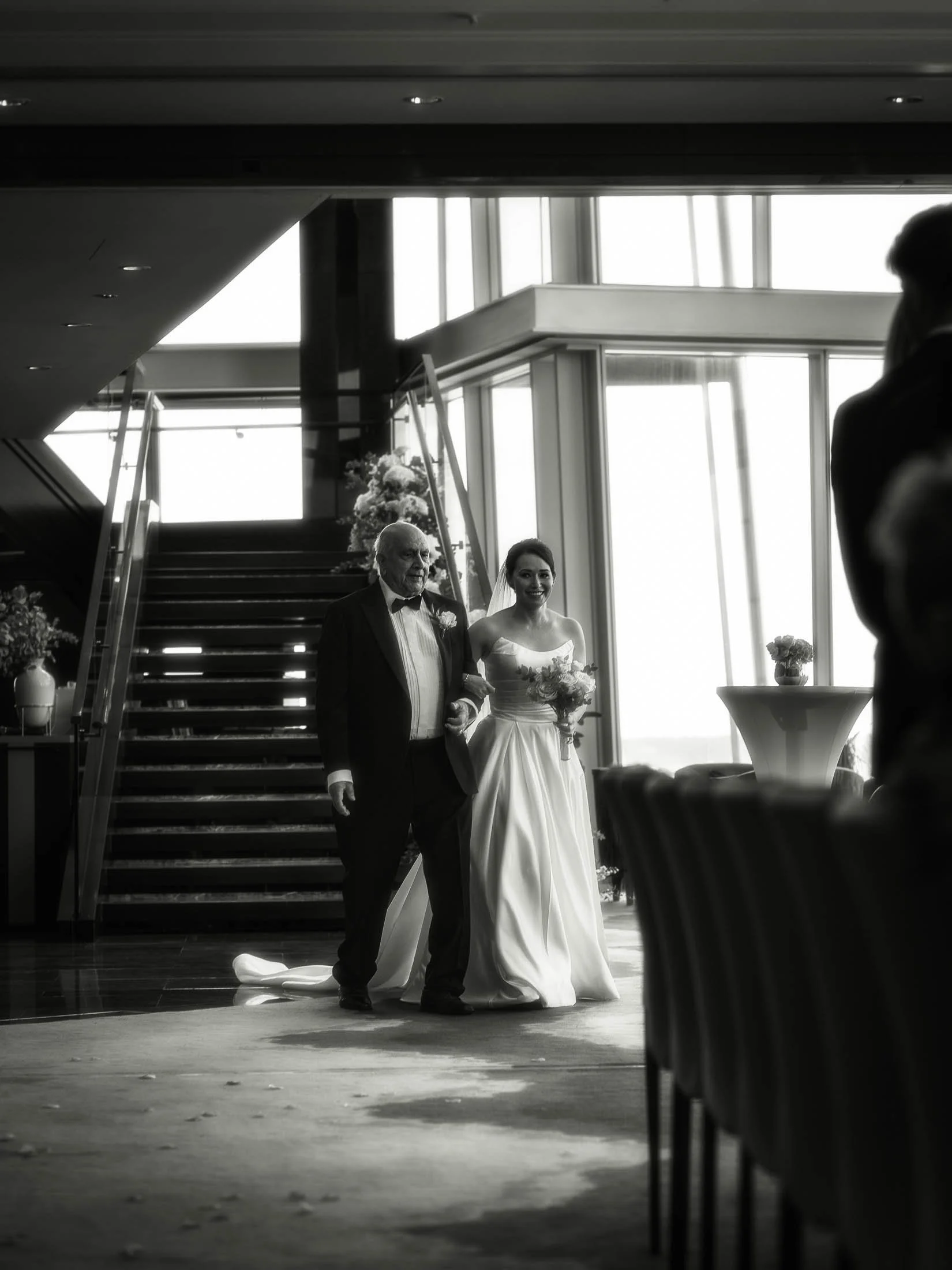 A black and white editorial wedding photograph of a bride walking arm-in-arm with her father into her wedding ceremony in London.