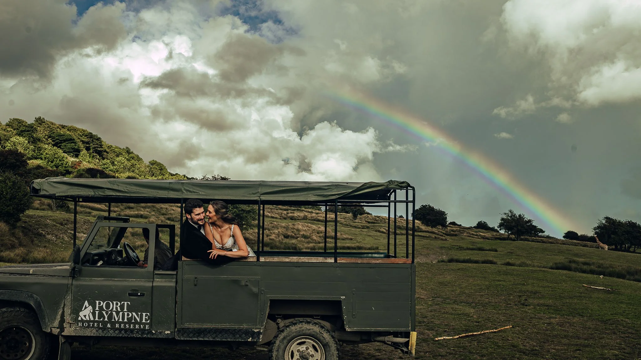 A couple laughing in a safari jeep at a Port Lympne wedding with a rainbow in the background
