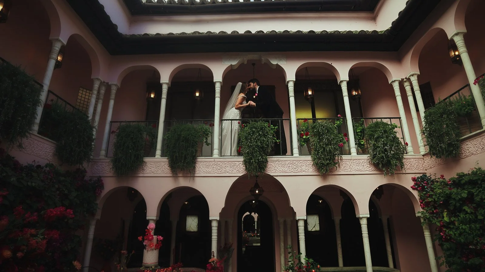 A wide shot of a bride and groom kissing on a balcony, surrounded by many archways in a courtyard. Framed with editorial intent.