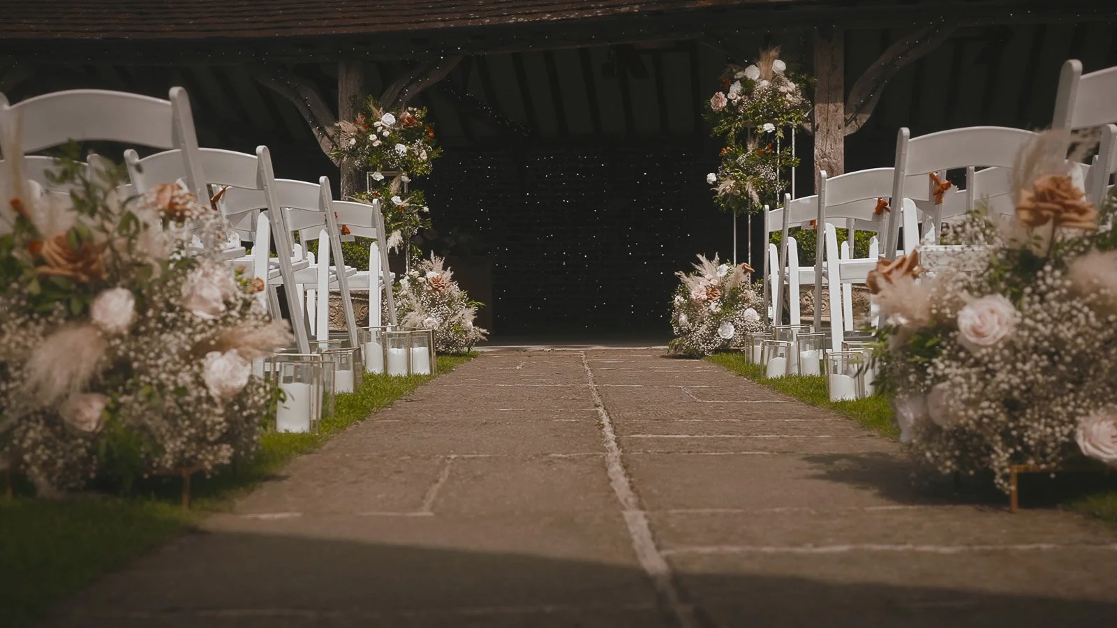 Outdoor ceremony aisle at Winters Barns with white chairs, meadow-style florals and glass candle lanterns in Kent.