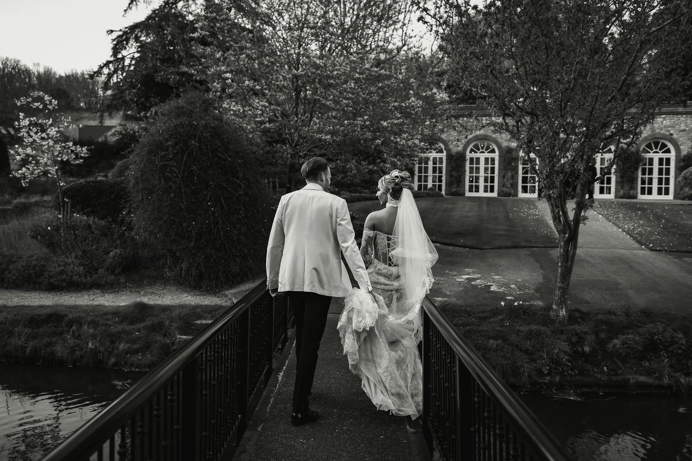 Editorial wedding moment as a groom in white tuxedo carries the dress of his bride across the bridge at The Orangery Maidstone.
