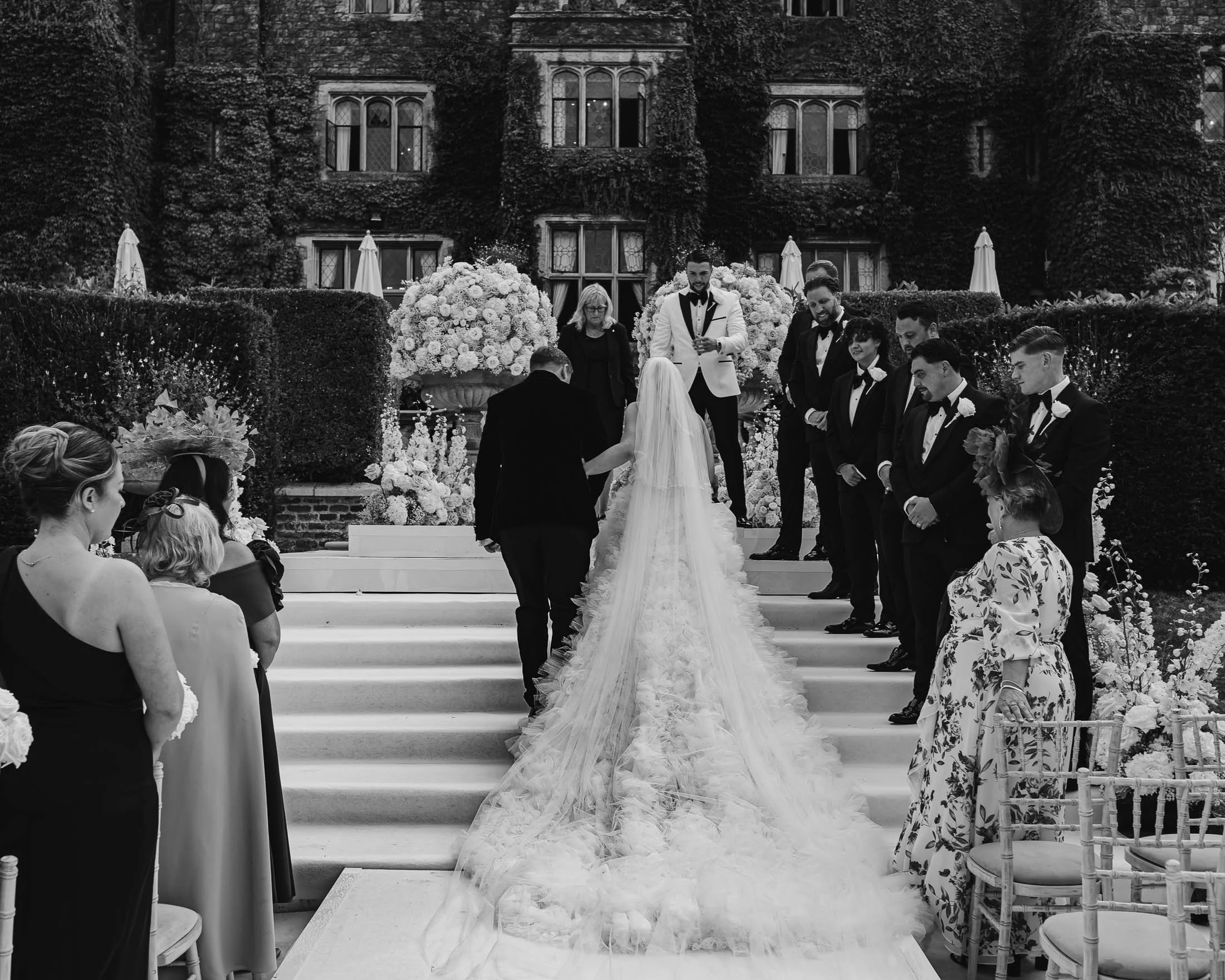 A brides long wedding dress train flows down white steps as she enters her outdoor ceremony in a black and white fine art wedding photography and videography moment.