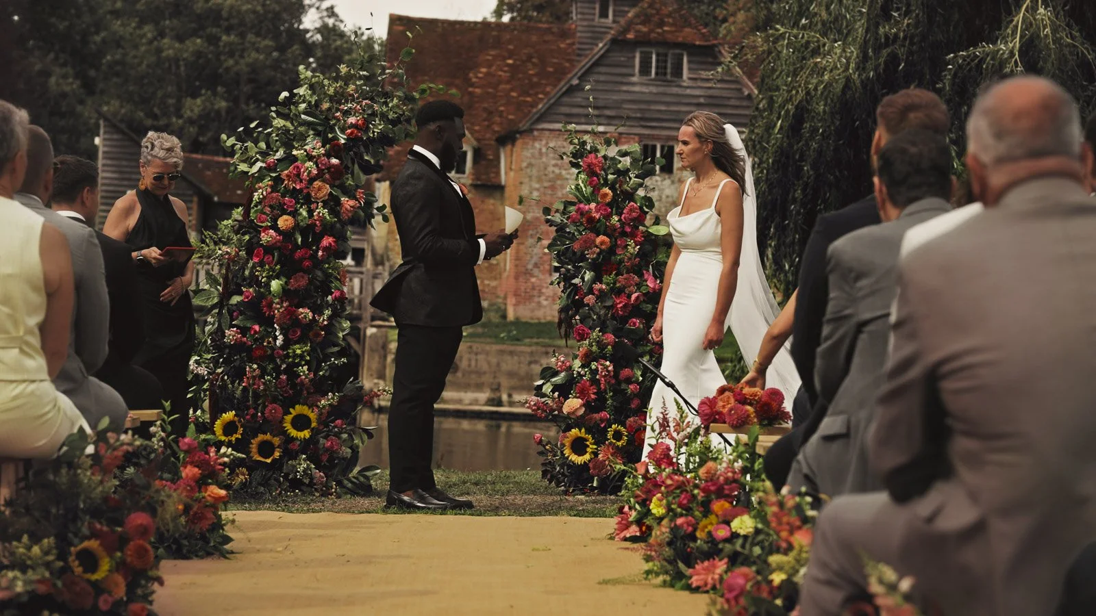An outdoor wedding ceremony by the River Thames during a colourful riverside wedding day. The groom is wearing black tie and reading his personal vows, whilst the bride is wearing a luxurious, simple gown.