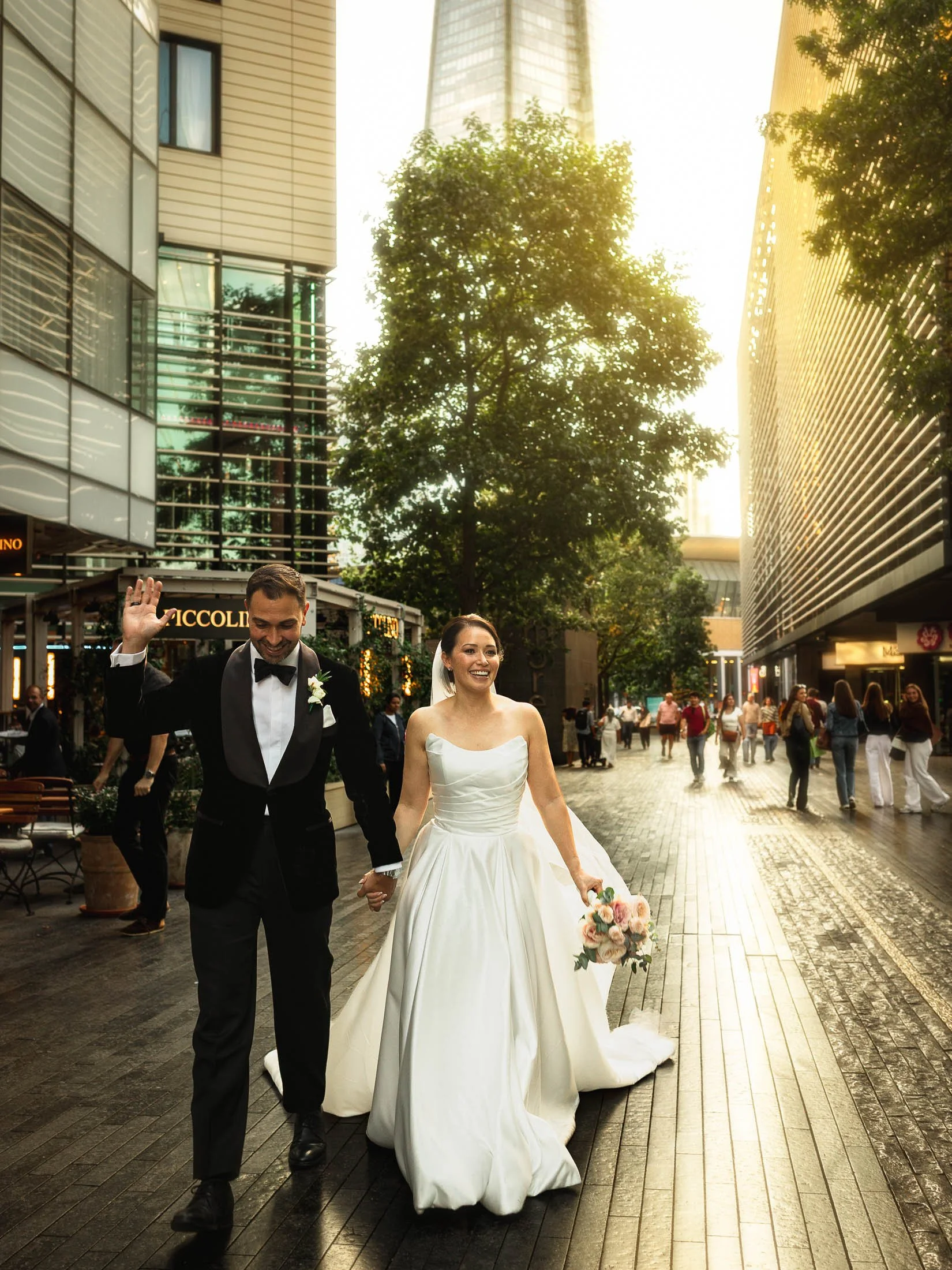 A groom in black tie tuxedo holds the hand of his bride and acknowledges passers by during their wedding portrait session on the streets of London during golden hour.