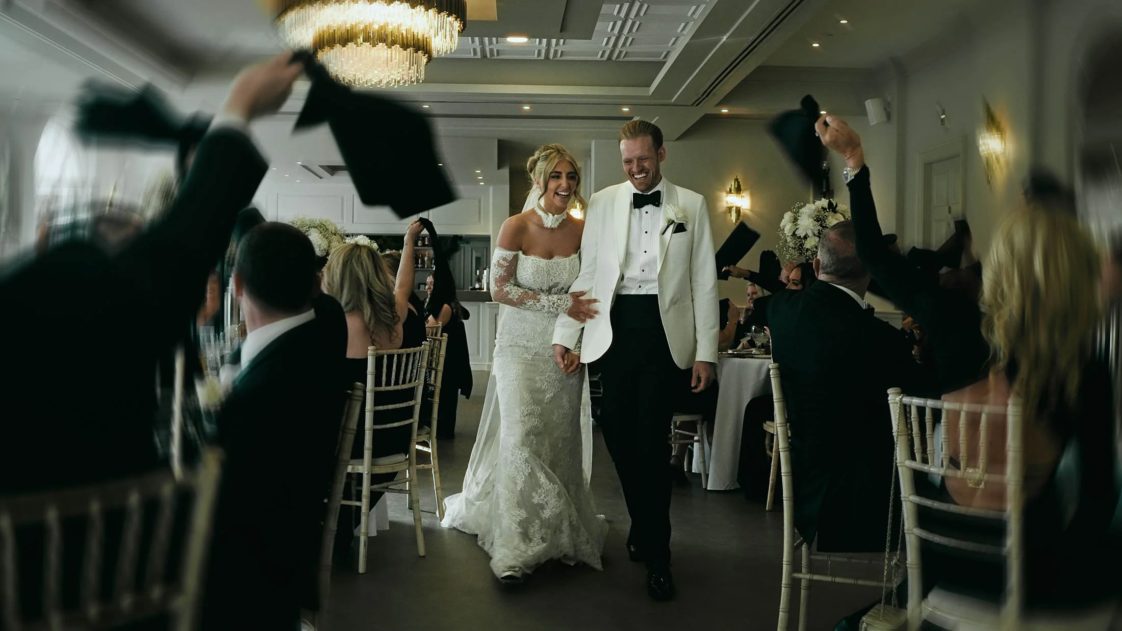 A couple laugh together as they are welcomed into their wedding reception by guests cheering, taken from their cinematic storytelling wedding film captured at The Orangery, Maidstone, in the UK.