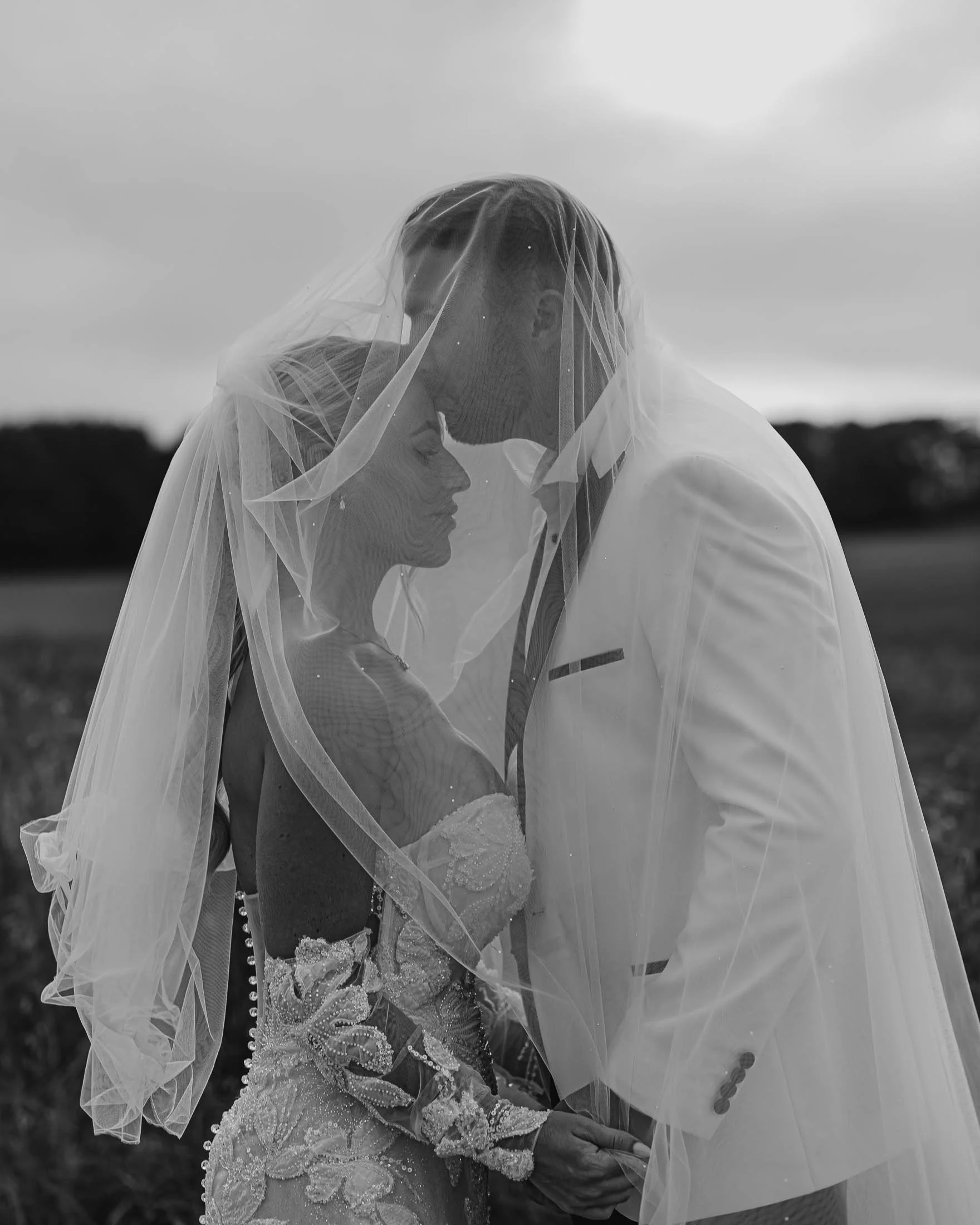 A black and white photograph of a bride and groom sharing a kiss under a veil, with the bride in an ornately decorated dress and the groom in a light-colored suit, outdoors with a blurred natural background.