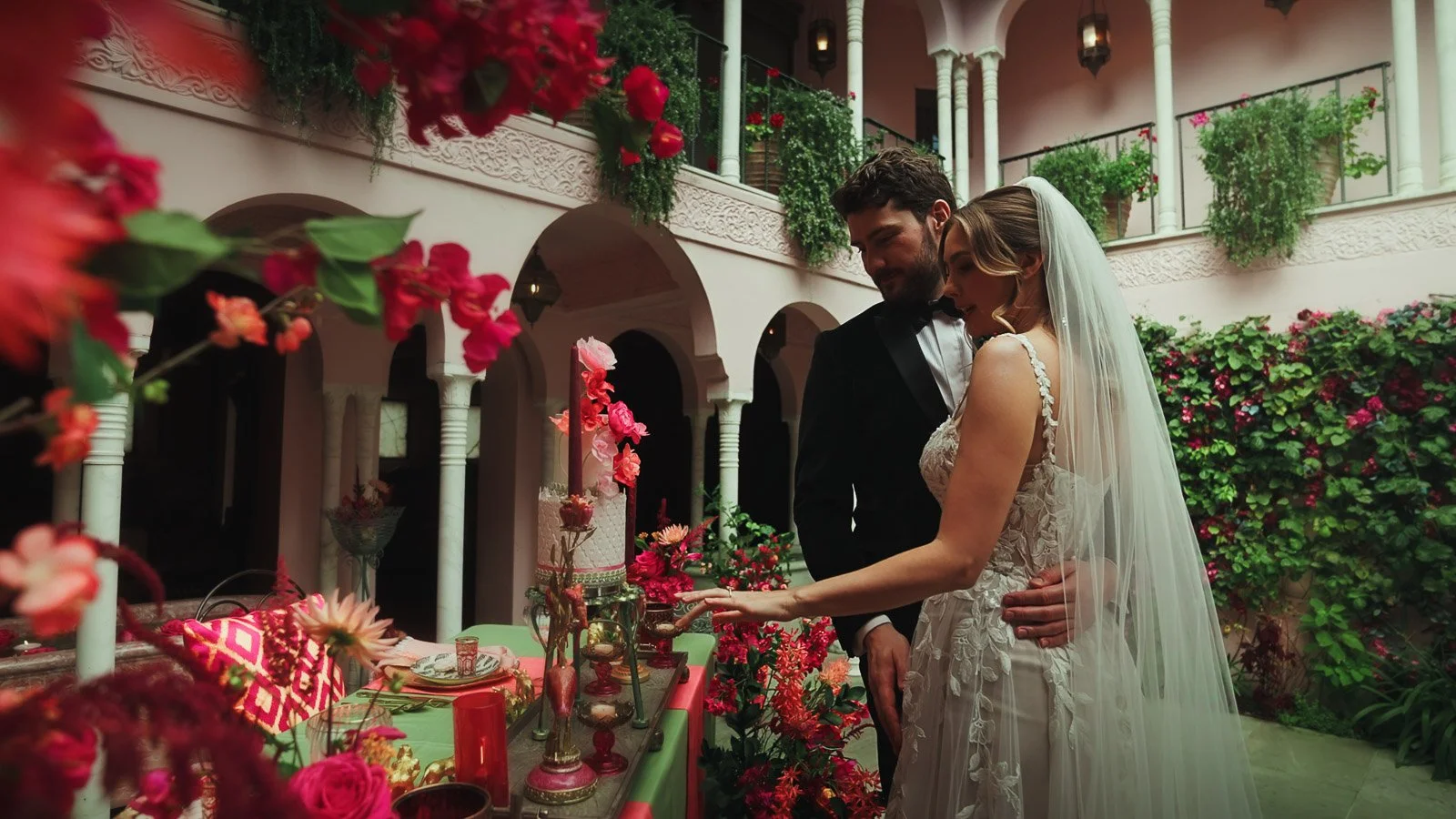 A couple cut their wedding cake in a courtyard, surrounded by bold pink and red florals and detailing during their black tie wedding.