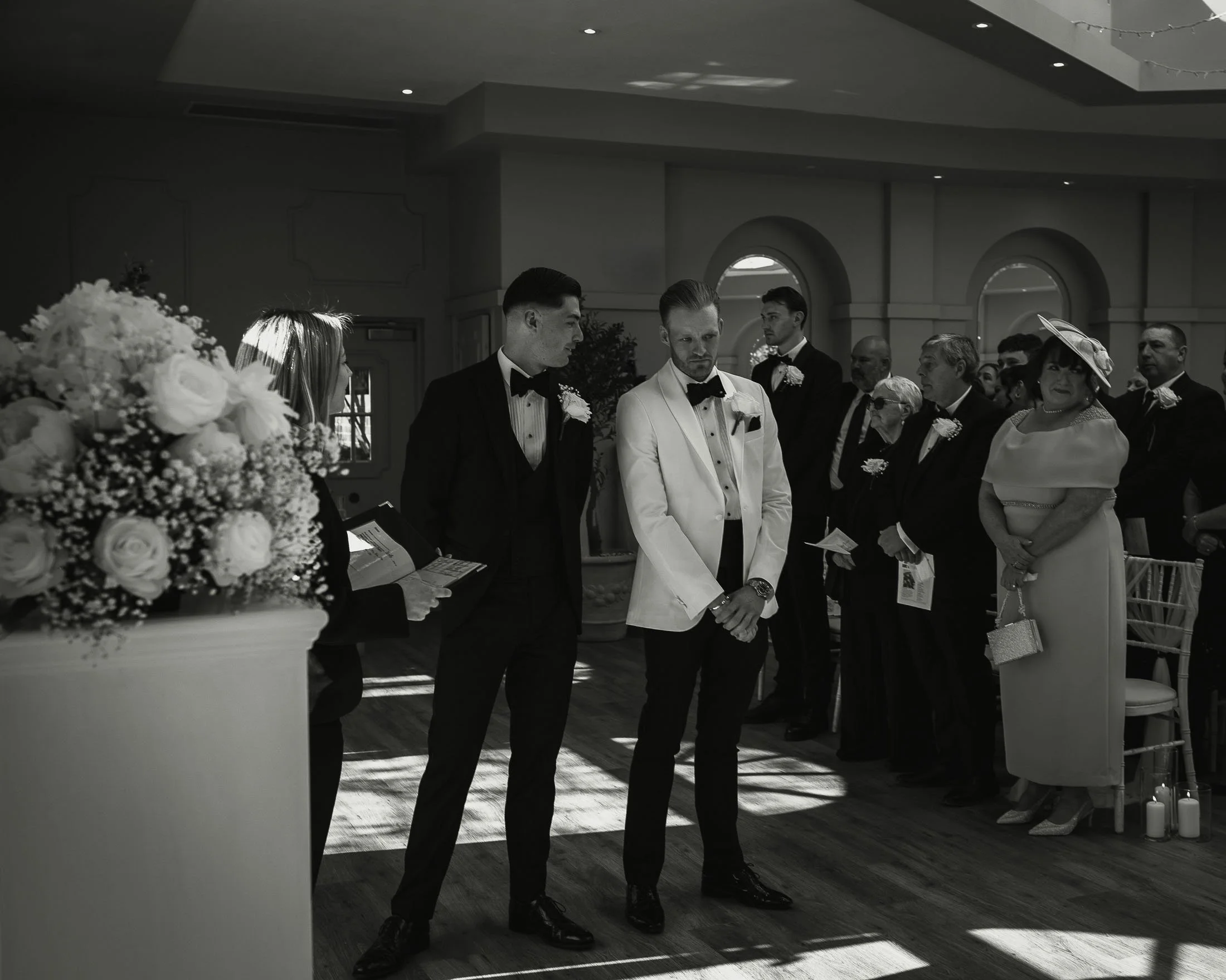 White tuxedo groom and black-tie best man wait at the end of the aisle during an indoor wedding ceremony in Kent.