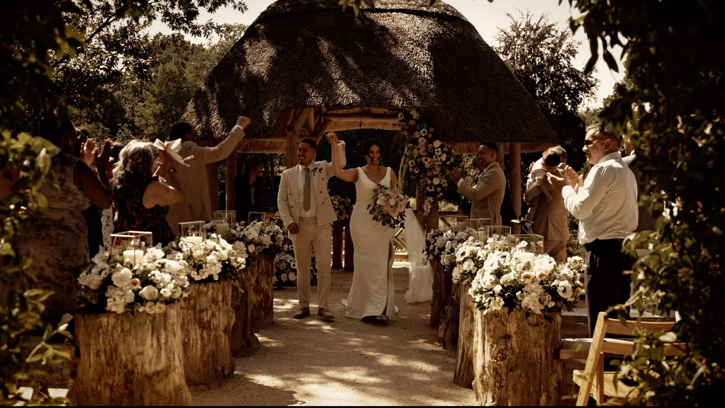 A bride and groom walking down the aisle after their outdoor summer wedding ceremony at  the Lapa at The Orangery, Maidstone in Kent, as guests cheer and throw confetti at them.