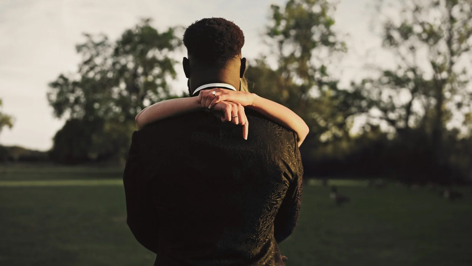 A man in a black suit holding a woman in a white dress in his arms during sunset in a park.
