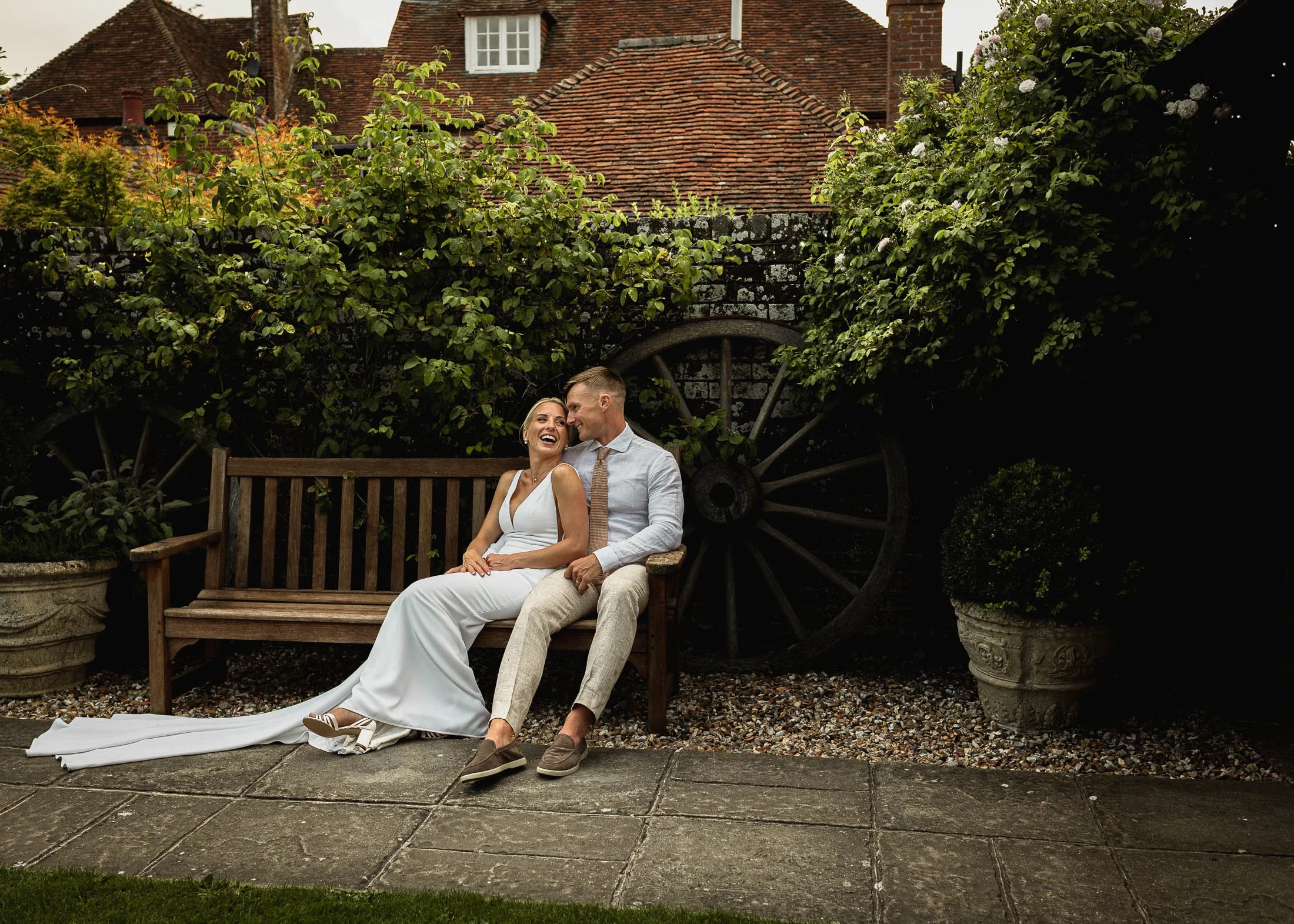 A bride and groom sit on a bench together and laugh during their portrait session in the courtyard of Winters Barns wedding venue.