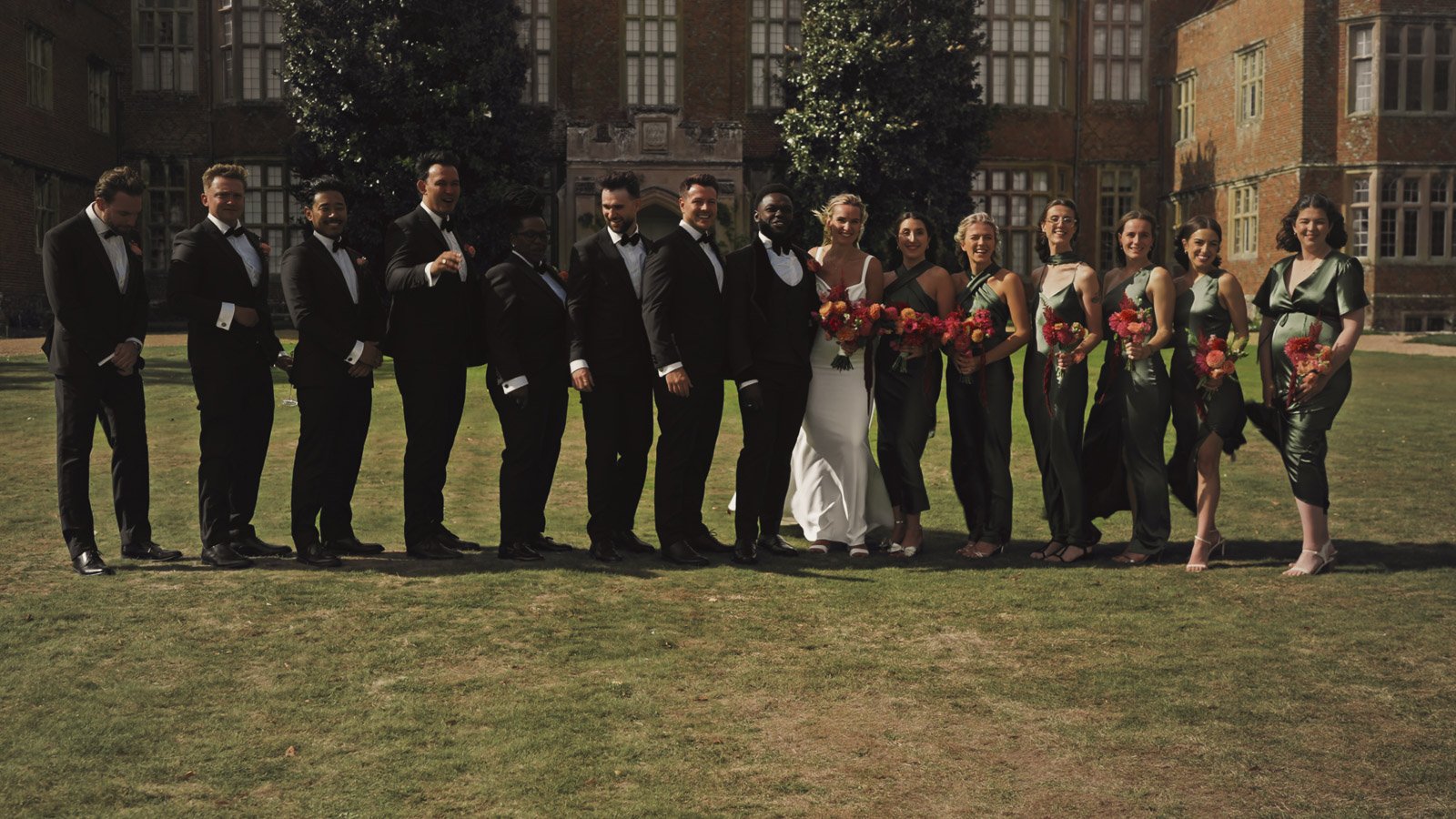 A bridal party wearing black tie and emerald green bridesmaids dresses pose for a group shot during a colourful riverside wedding at an Oxfordshire Estate.