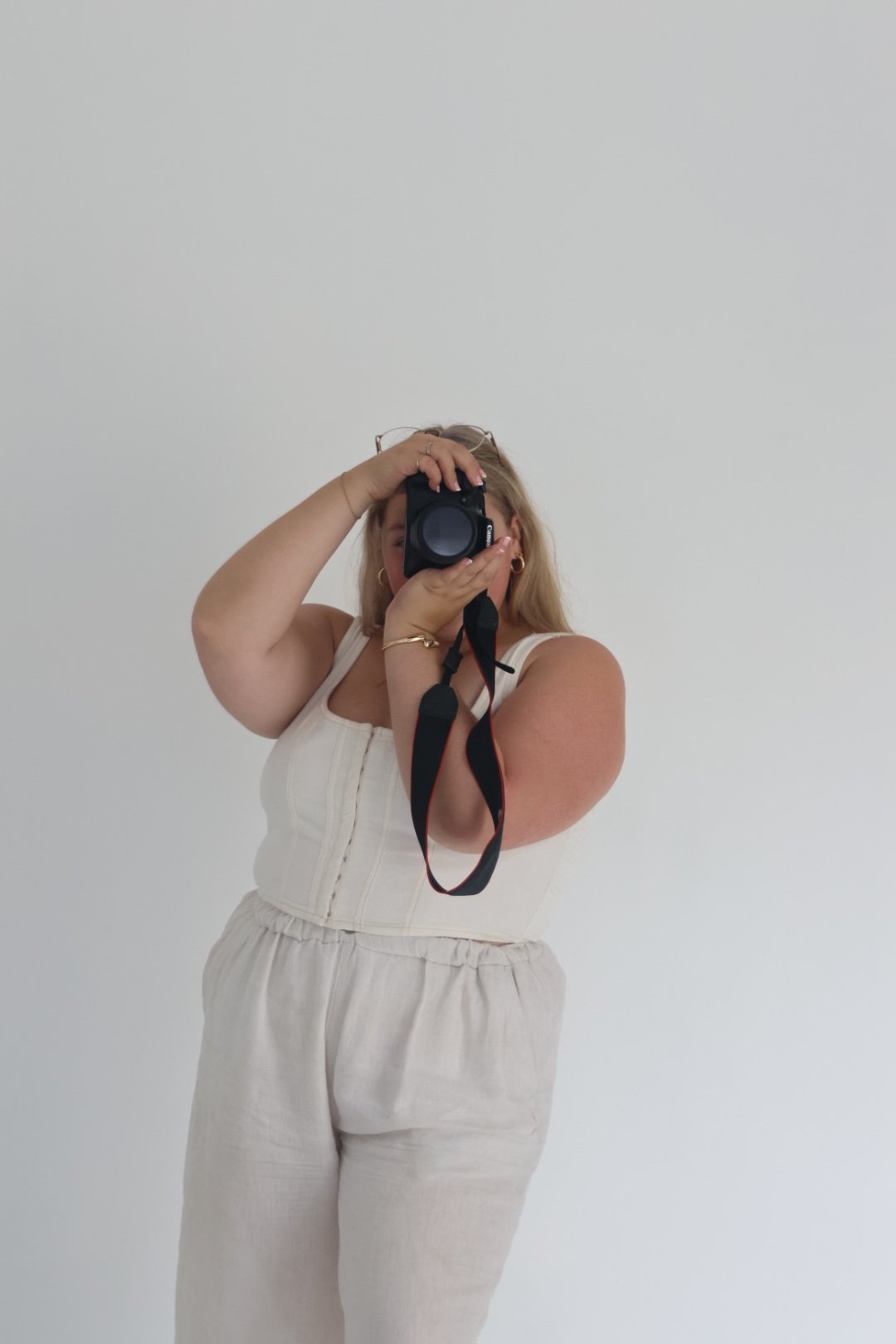 A woman with blonde hair taking a photo of herself with a camera, standing against a plain white wall.